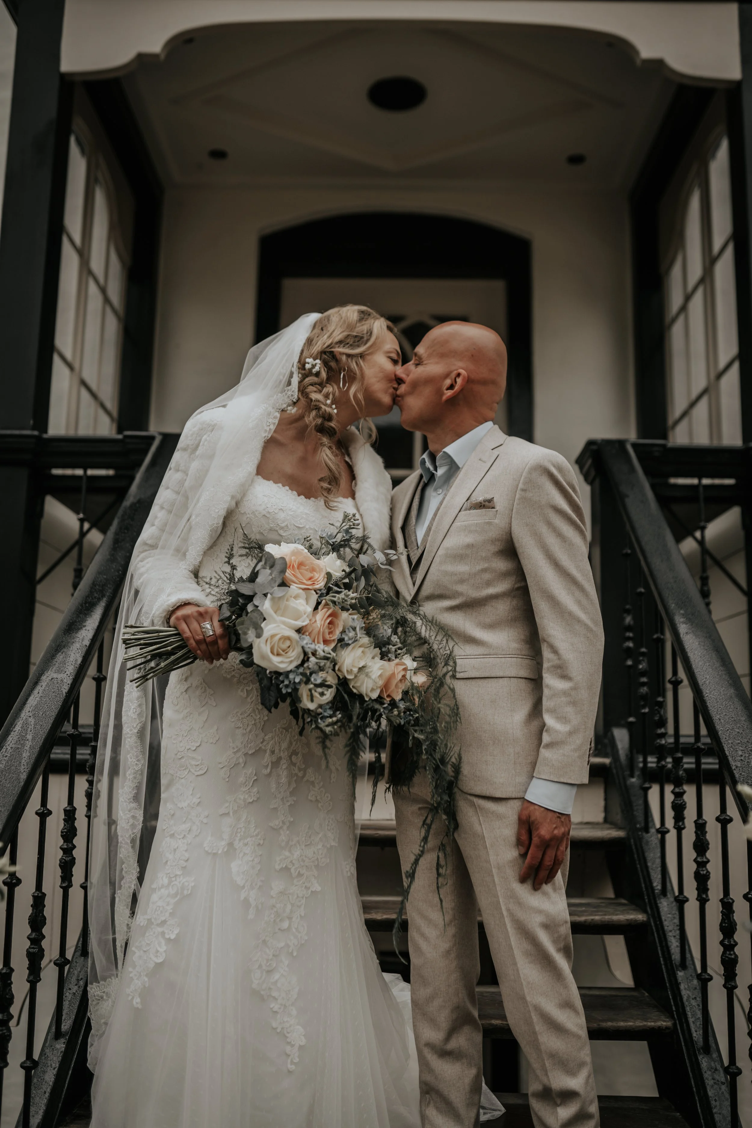 Een bruid en bruidegom kussen elkaar op een trap voor een oud gebouw. De bruid houdt een bos bloemen.