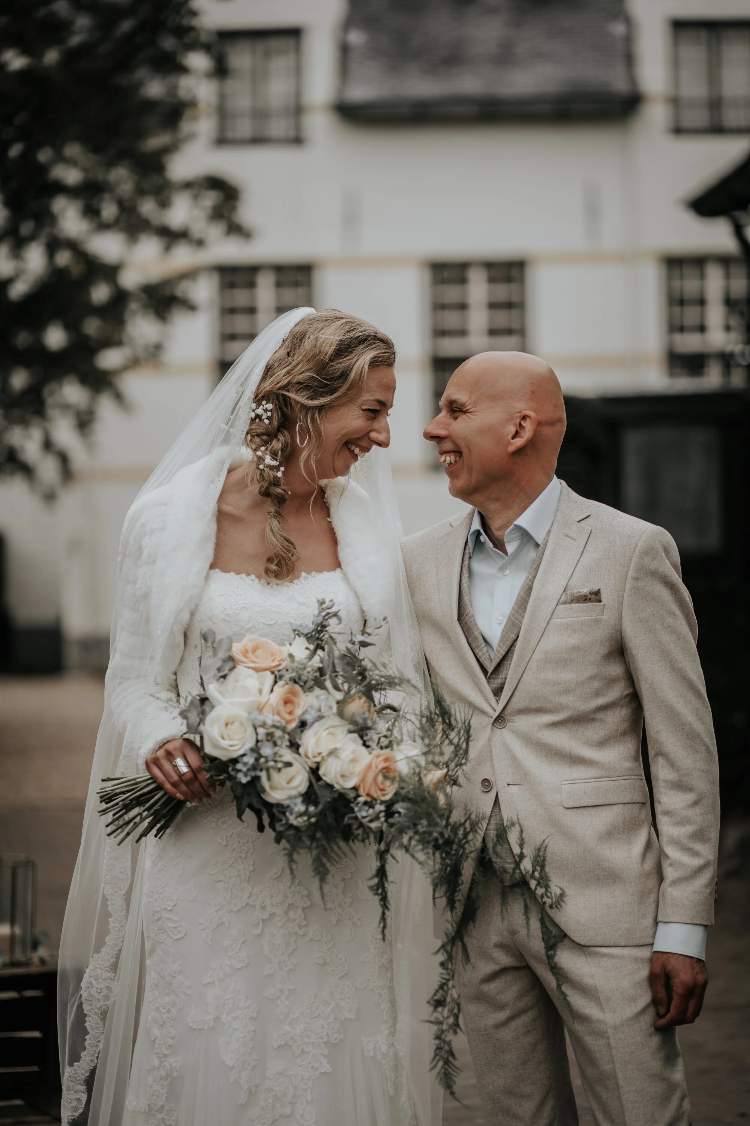 Een bruid en een bruidegom die lachen naar elkaar, buiten op de bruiloft, met de bruid in een witte jurk en een sluier, en de bruidegom in een beige kostuum.