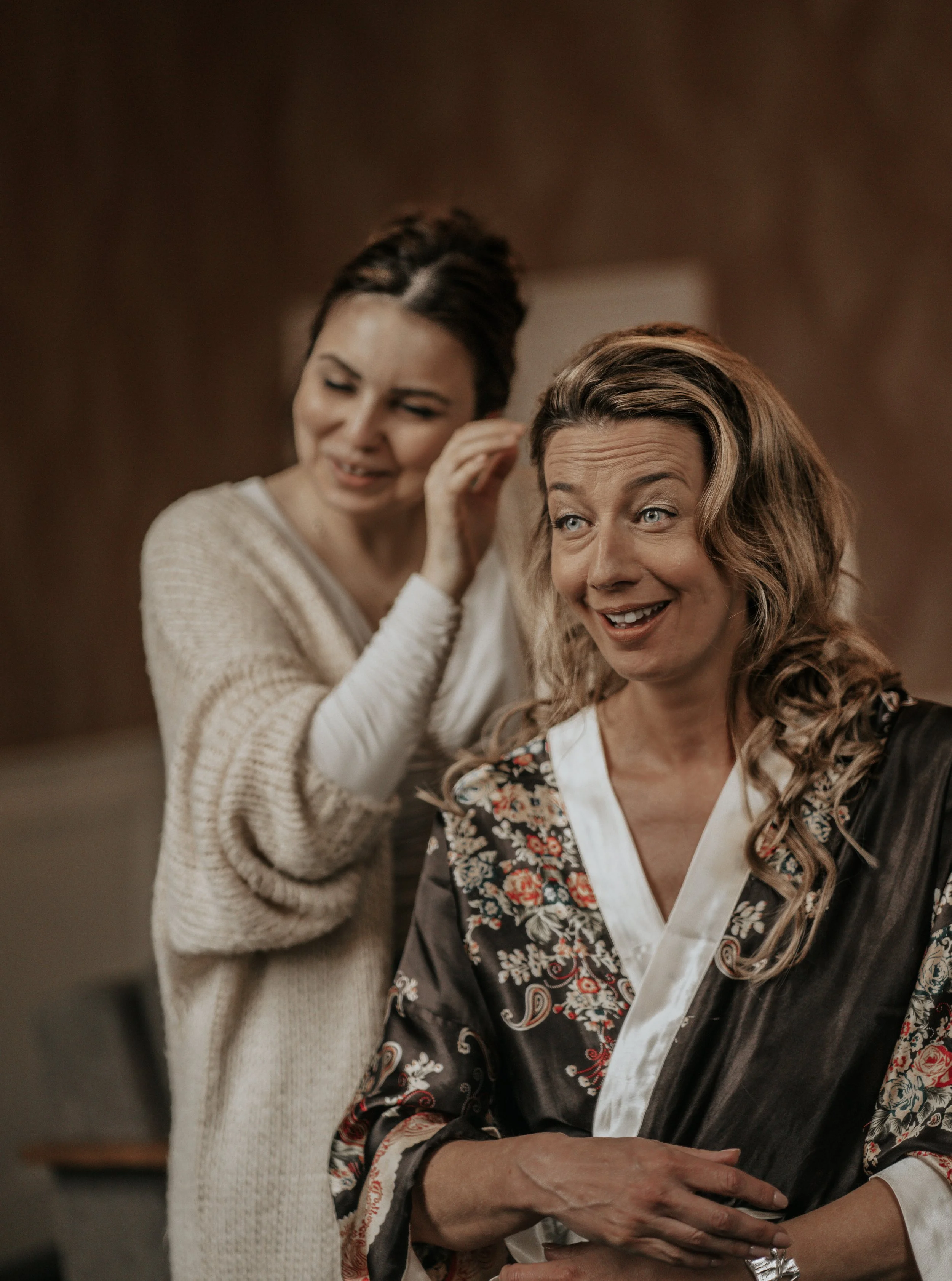 Twee vrouwen, een met lichtbruin haar en een met donker haar, zitten in een kamer en lijken vrolijk te zijn. De vrouw met lichtbruin haar draagt een zwarte zijden kimono met bloemen, terwijl de andere vrouw een beige gebreide trui draagt.