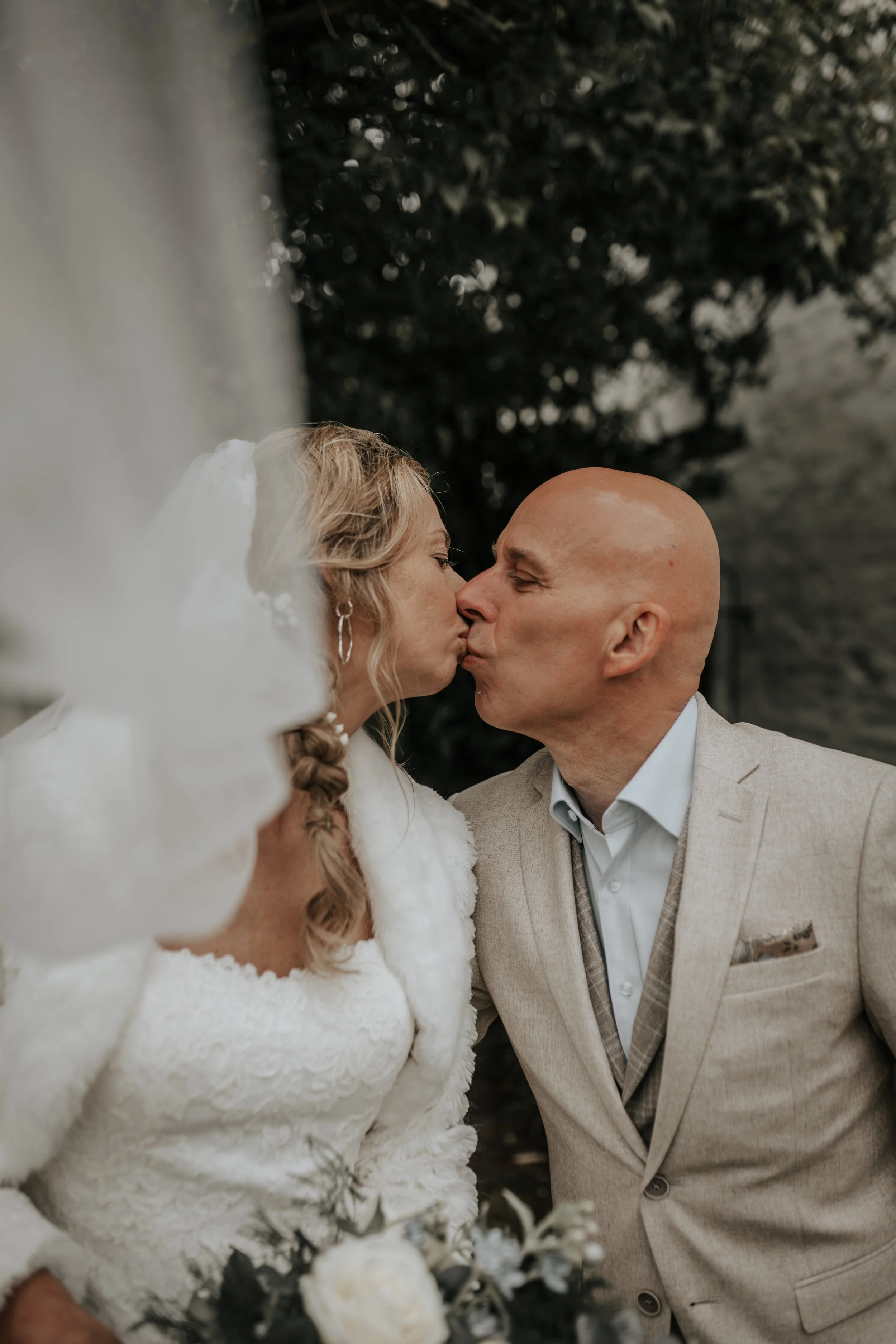 Een stel kust elkaar tijdens een bruiloft, de vrouw draagt een witte jurk en een witte jas, de man een beige pak. Ze staan buiten met een donkere achtergrond van bomen.