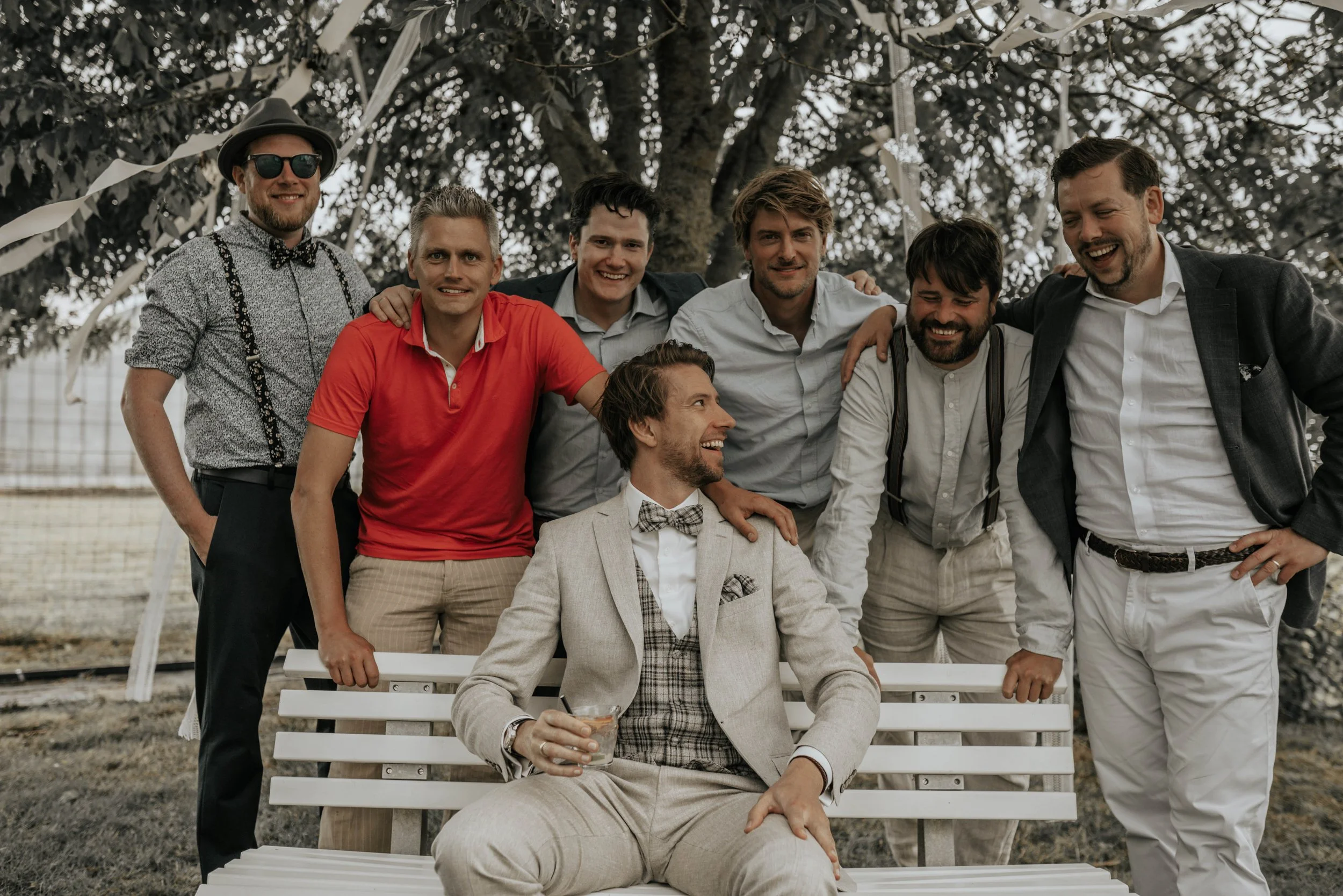 Vijftien jonge mannen die samen lachen en poseren in een tuin onder een boom, met één man in een beige pak zittend op een bank en de rest staand eromheen.