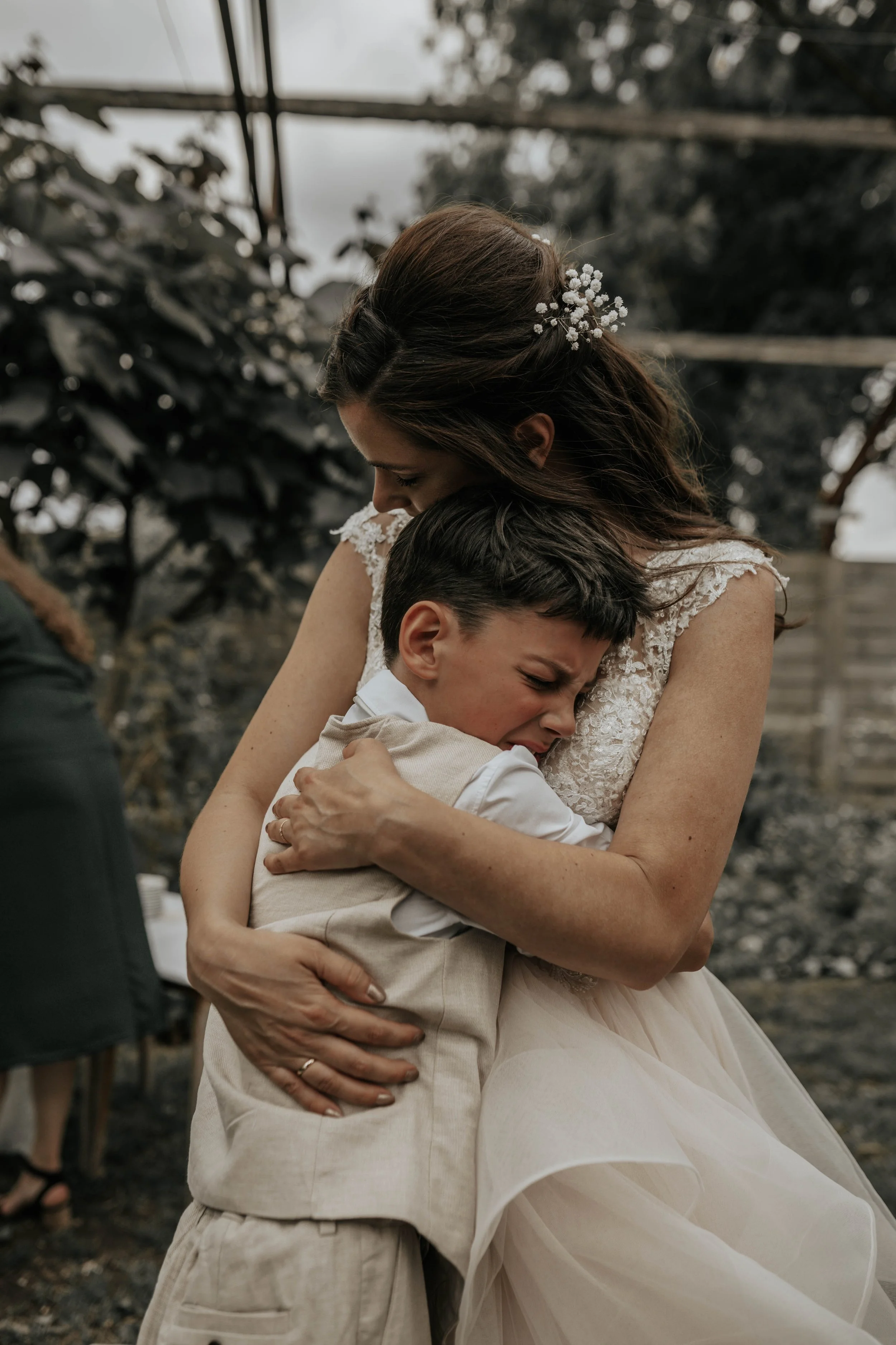 Een vrouw en een jongen omhelzen elkaar emotioneel, mogelijk tijdens een bruiloft, binnen in een kas of serre met planten op de achtergrond.