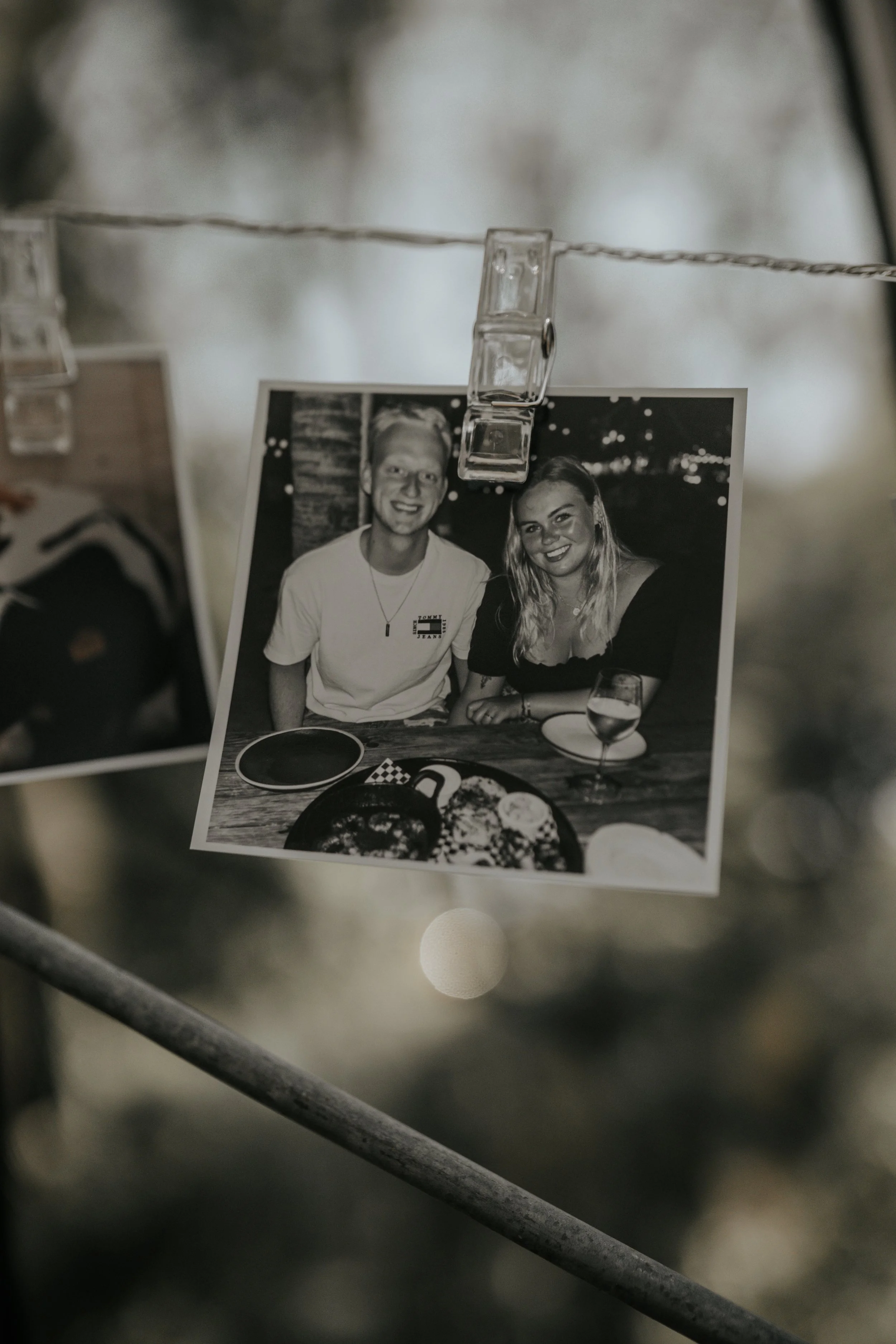 Zwart-wit foto van een lachend stel dat samen aan een tafel zit met eten en drinken, opgehangen aan een lijn met een knijper.