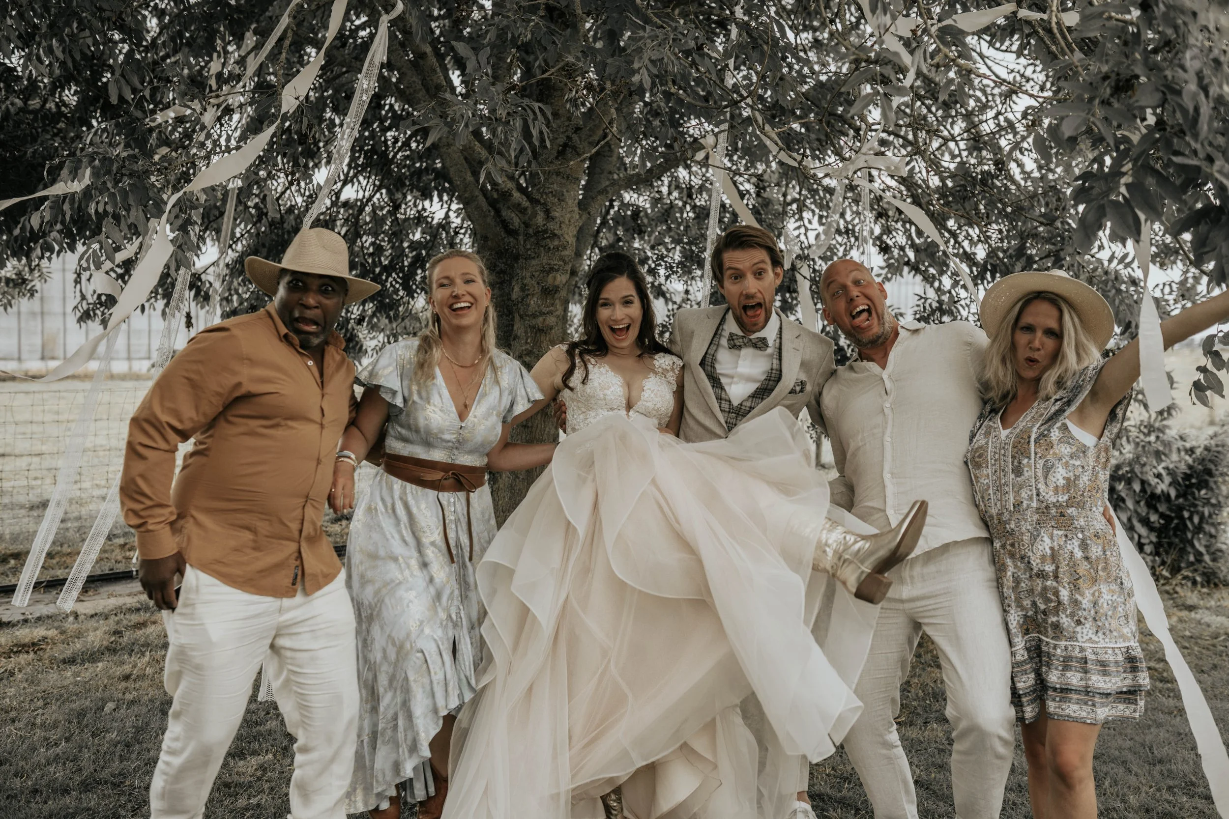 Mensen vieren een bruiloft onder een boom, lachen en poseren voor de foto.