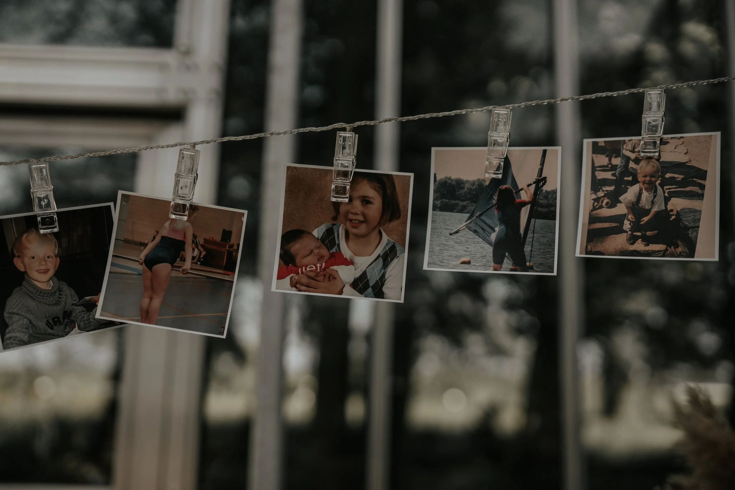 Vijf foto's hangen aan een draad, vastgeklikt met transparante klemmen. Ze tonen kinderen die binnen en buiten spelen, een meisje dat met een baby knuffelt, en een kind dat met een zeilboot op een rivier zeilt.
