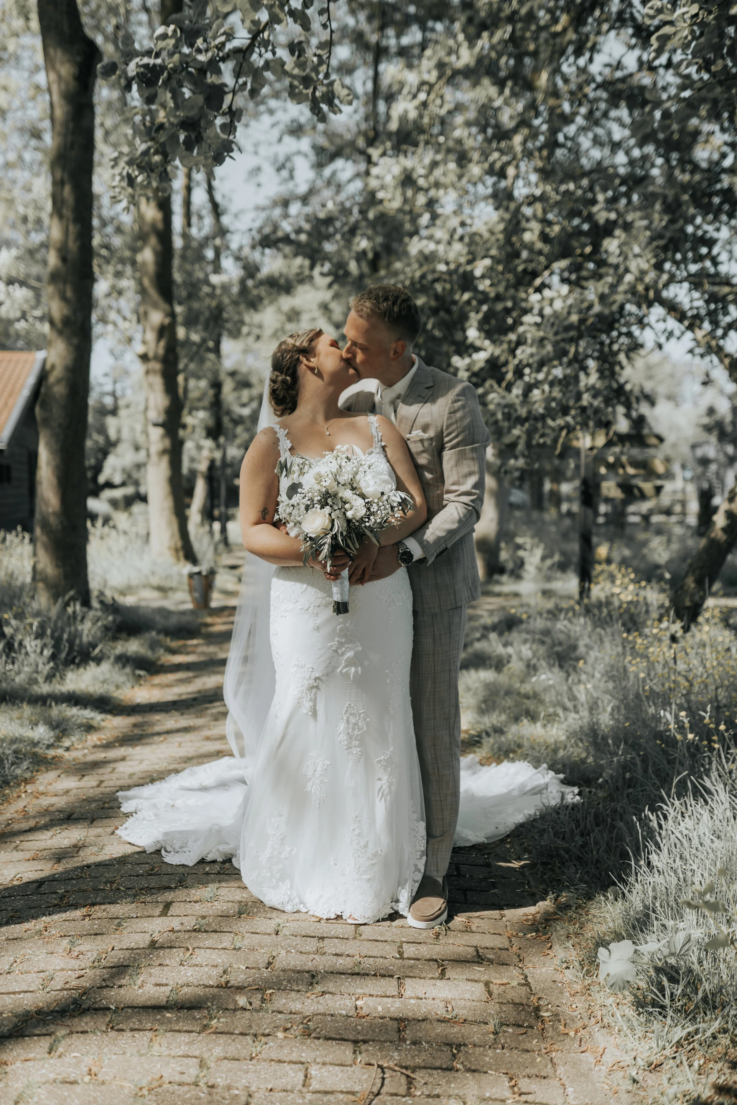 Een getrouwd stel staat op een houten pad in een park, de bruid houdt een bloementrouwstuk vast, ze kussen elkaar onder een boom op een zonnige dag.