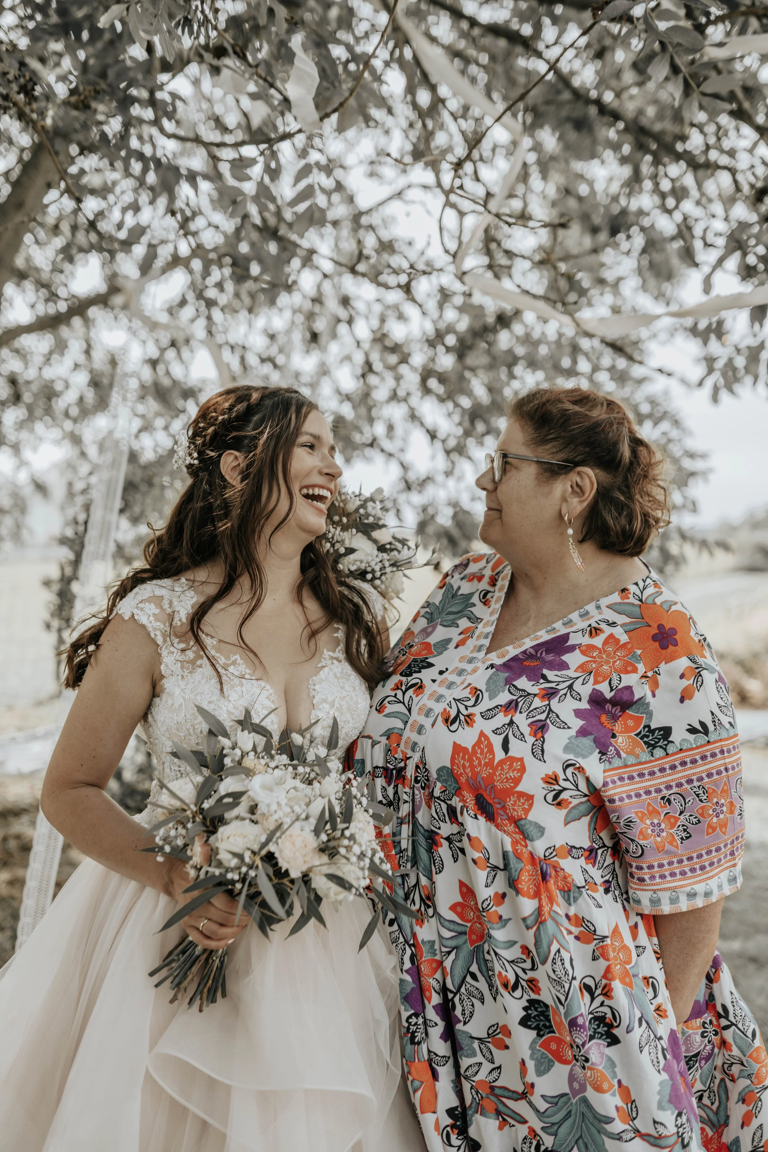 Vrouw in een trouwjurk met bloemen en een vrouw in een bloemenprint jurk die lachen naar elkaar, onder een boom.
