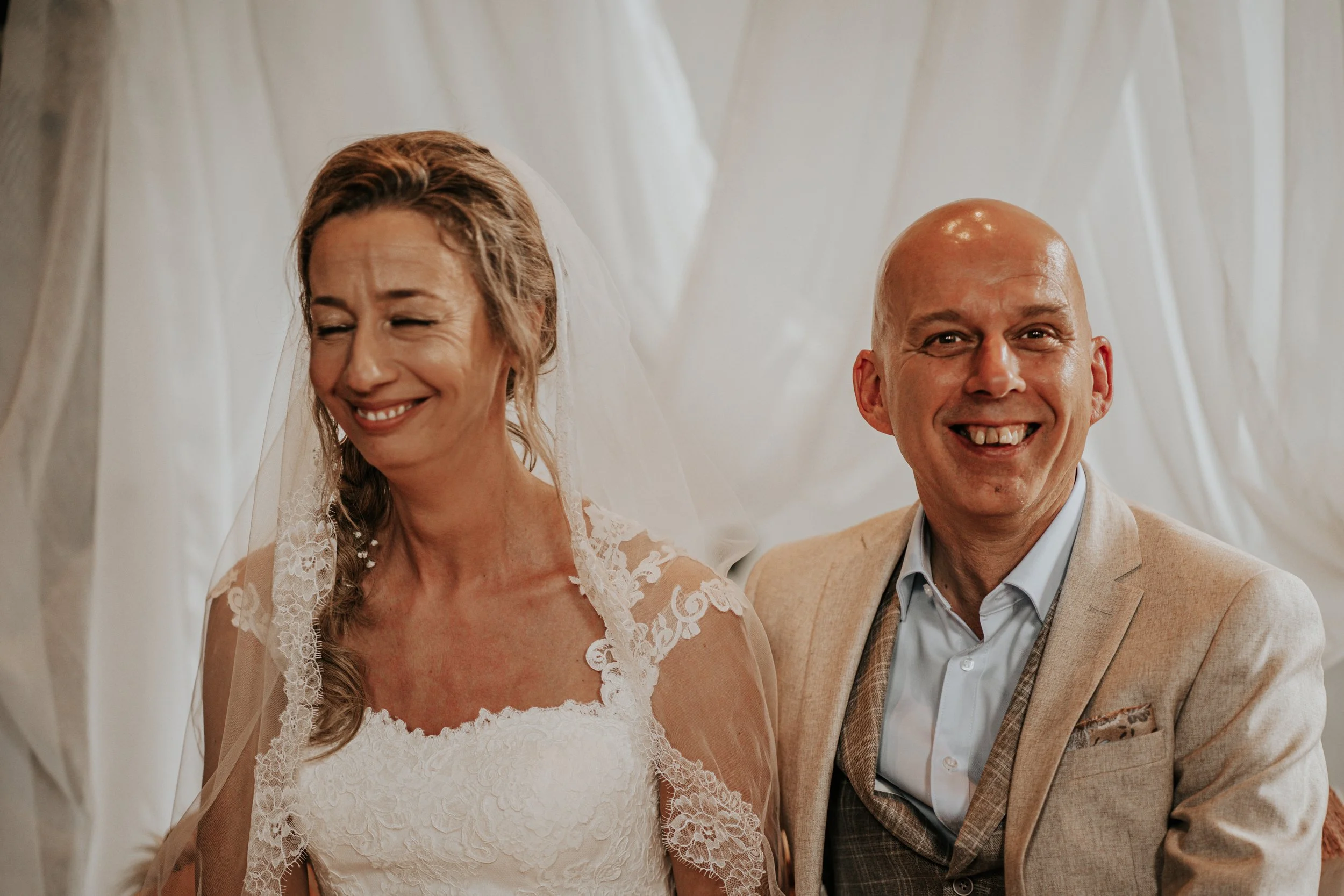 Een bruid en een bruidegom zitten samen, de bruid glimlacht met haar ogen dicht, de bruidegom lacht breed en kijkt recht naar de camera, ze zitten binnen onder een witte tent of doek.