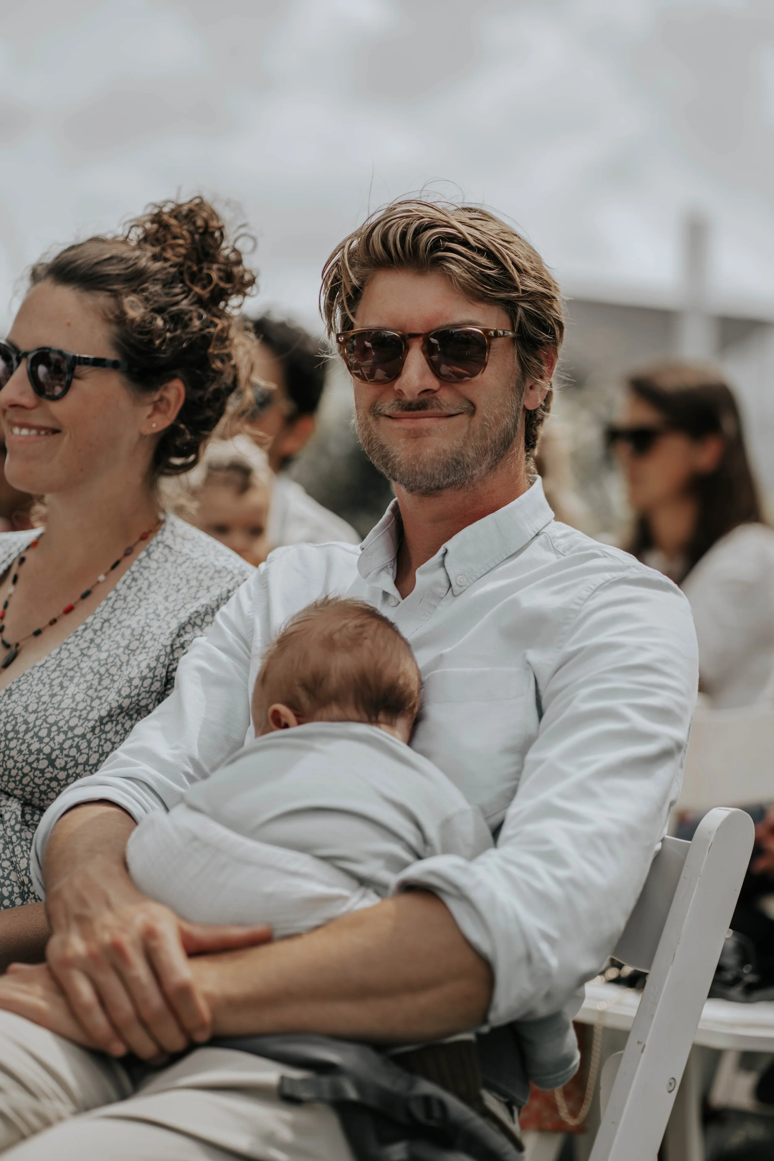 Een man met een witte blouse, zonnebril en een baby in zijn armen, zittend op een wit stoel tijdens een buitengebeurtenis, met meerdere mensen op de achtergrond.