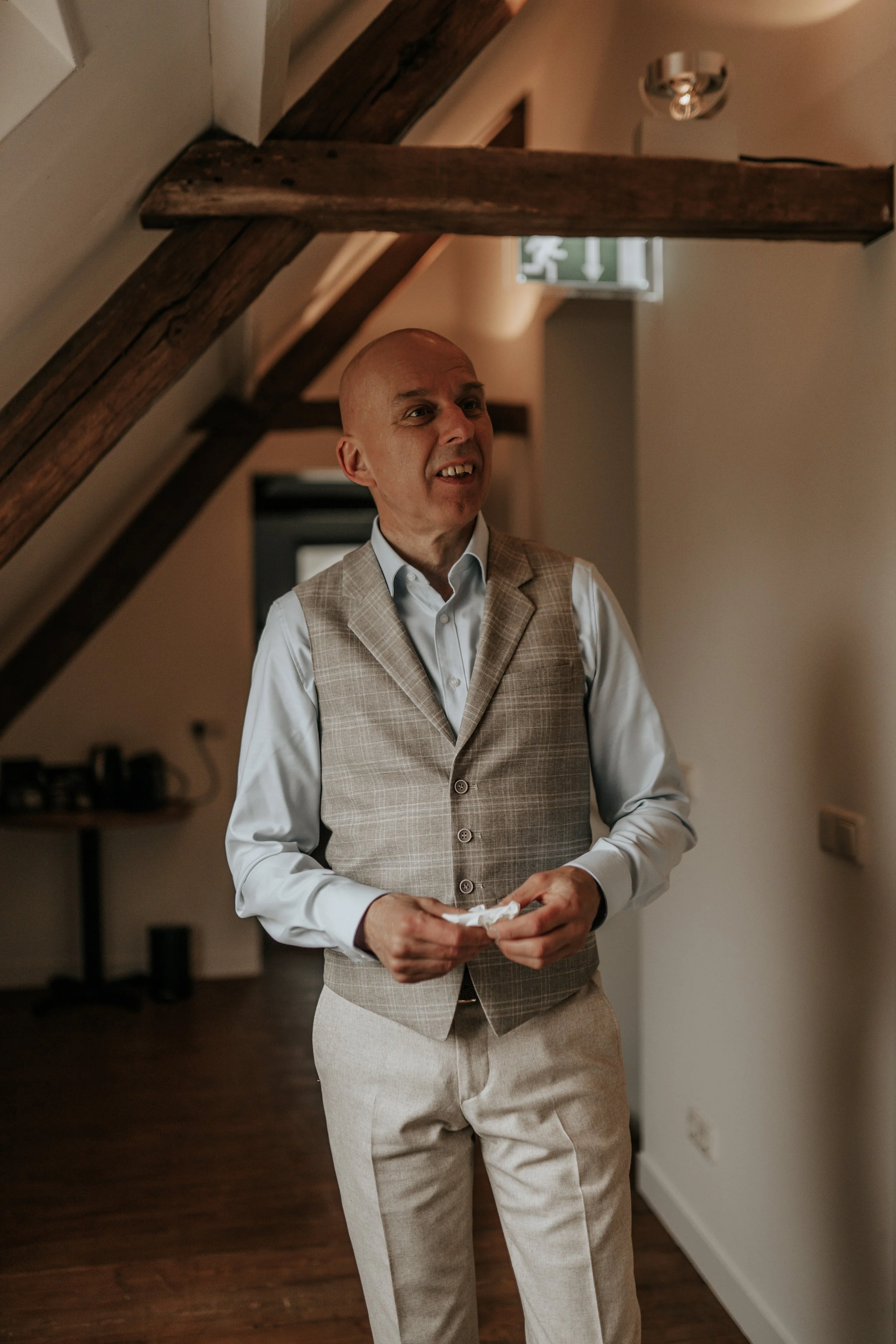 Man in formele kleding staat binnen in een huis, onder een houten steunbalk, met een uitdrukking van verwarring of verbazing.