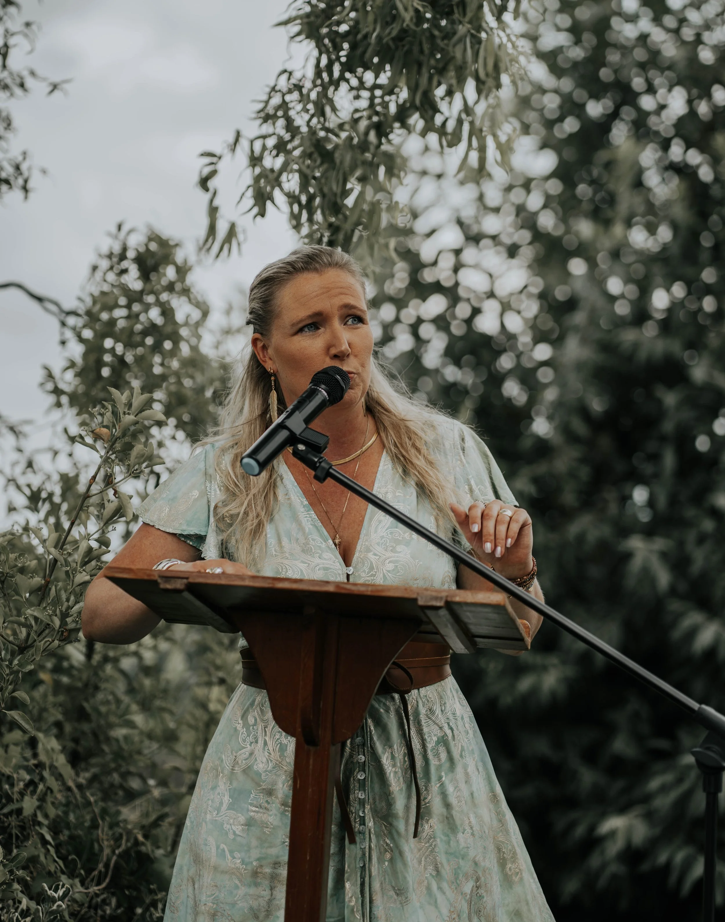 Vrouw staat buiten achter een houten lessenaar en spreekt in een microfoon, omringd door groene bladeren en bomen.