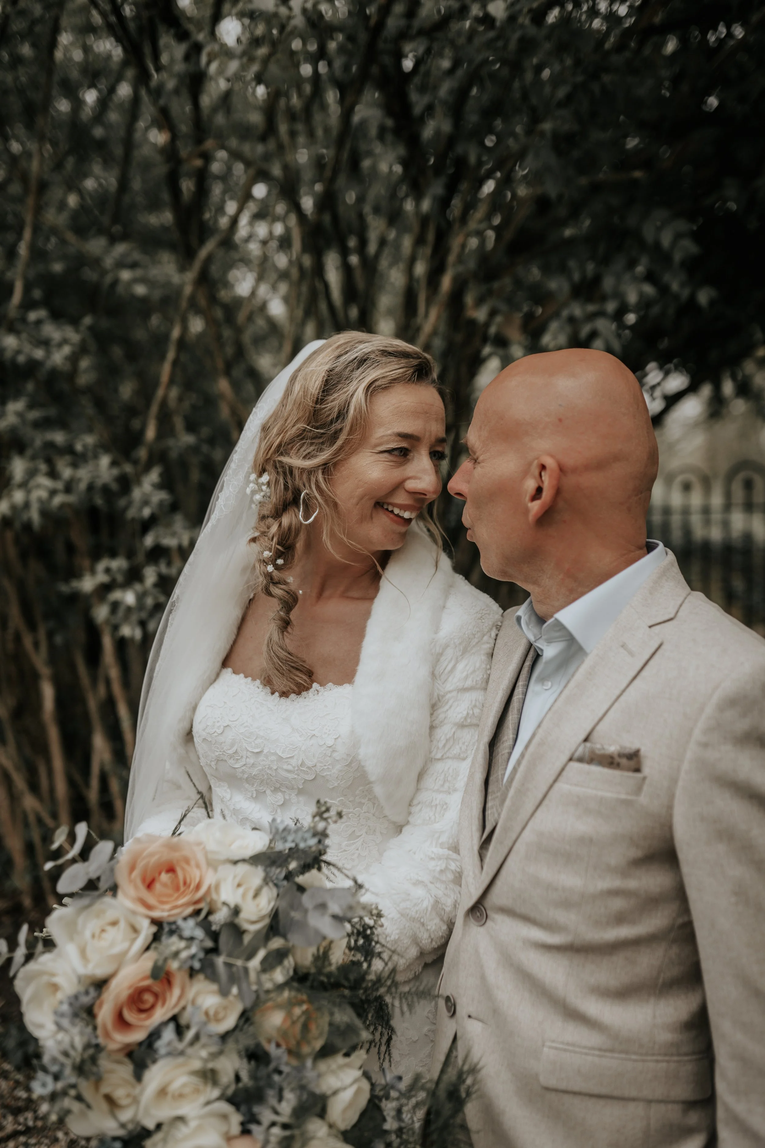 Een bruid en bruidegom die dicht bij elkaar staan, lachend en liefdevol naar elkaar kijken, buiten omgeven door bomen, met de bruid die een bloemenboeket vasthoudt.