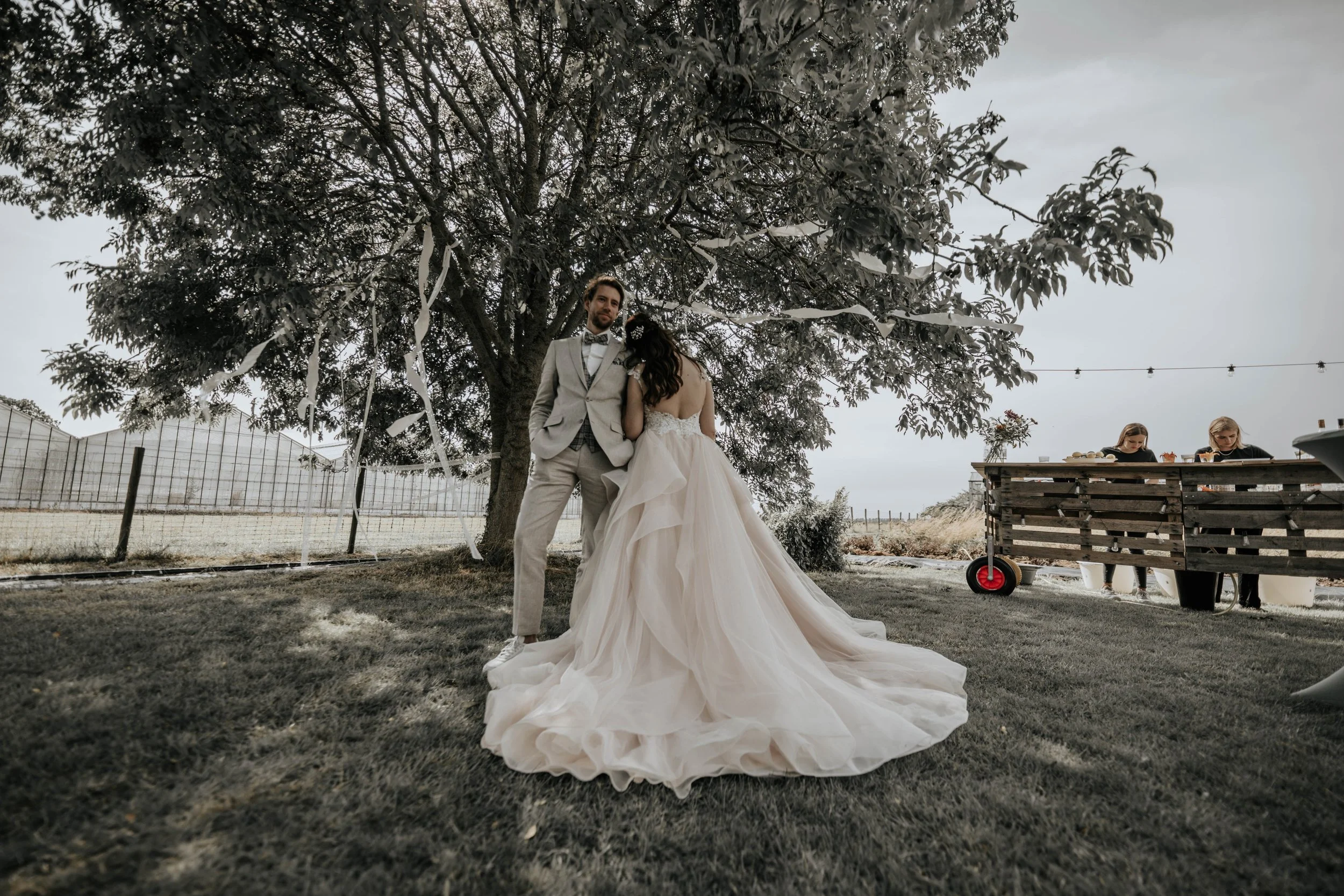 Een bruid en bruidegom staan onder een grote boom, dansend tijdens een bruiloft buiten. De bruid draagt een witte jurk met een lange sleep, de bruidegom draagt een lichtgrijs pak.
