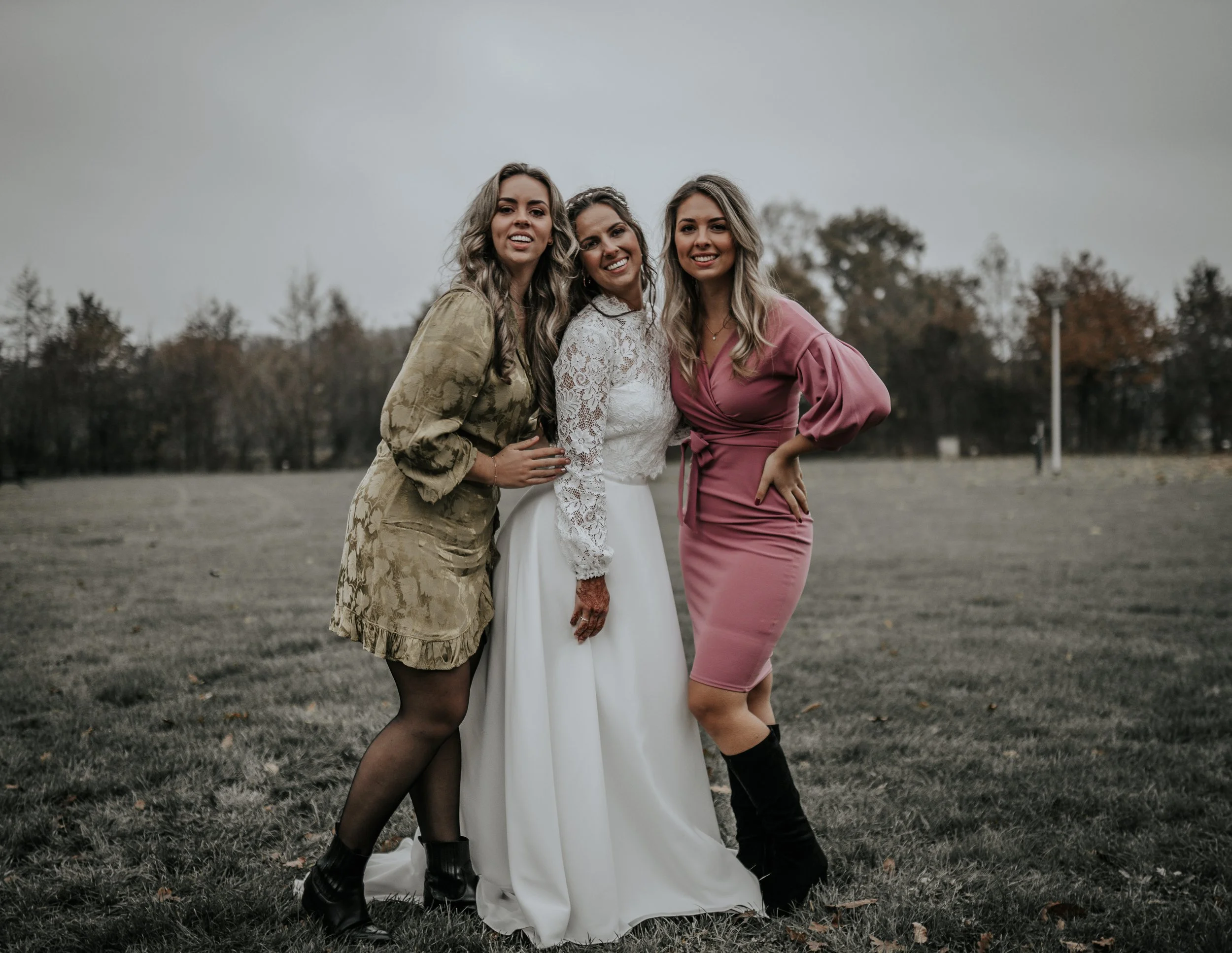 Drei vrouwen poseren in een veld, één draagt een witte trouwjurk, de andere twee dragen geklede outfits.
