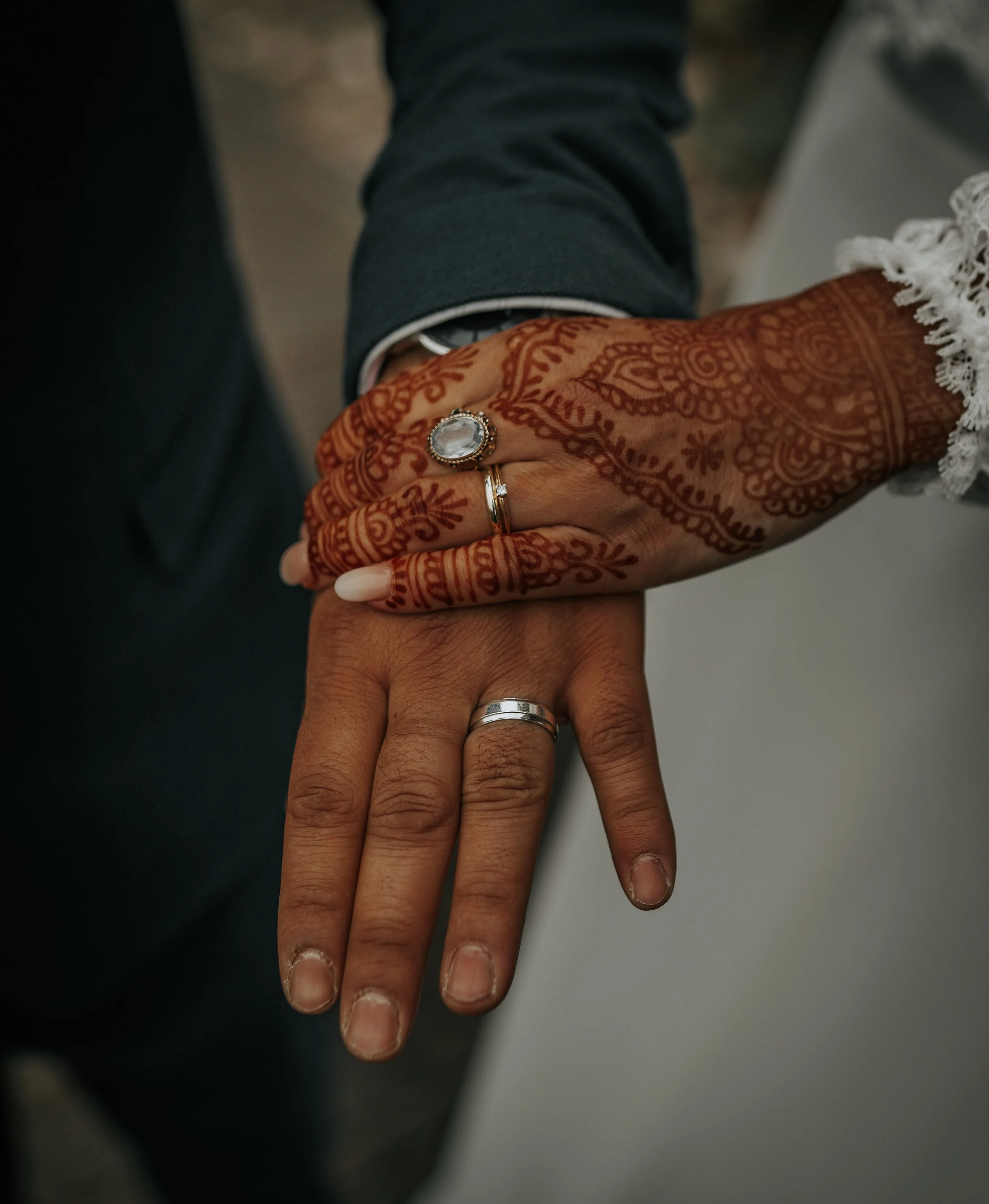 Handen van een getrouwde man en vrouw met trouwringen, de vrouw heeft een henna-tatoeage op haar hand en draagt een grote ring met een lichtgekleurde steen.