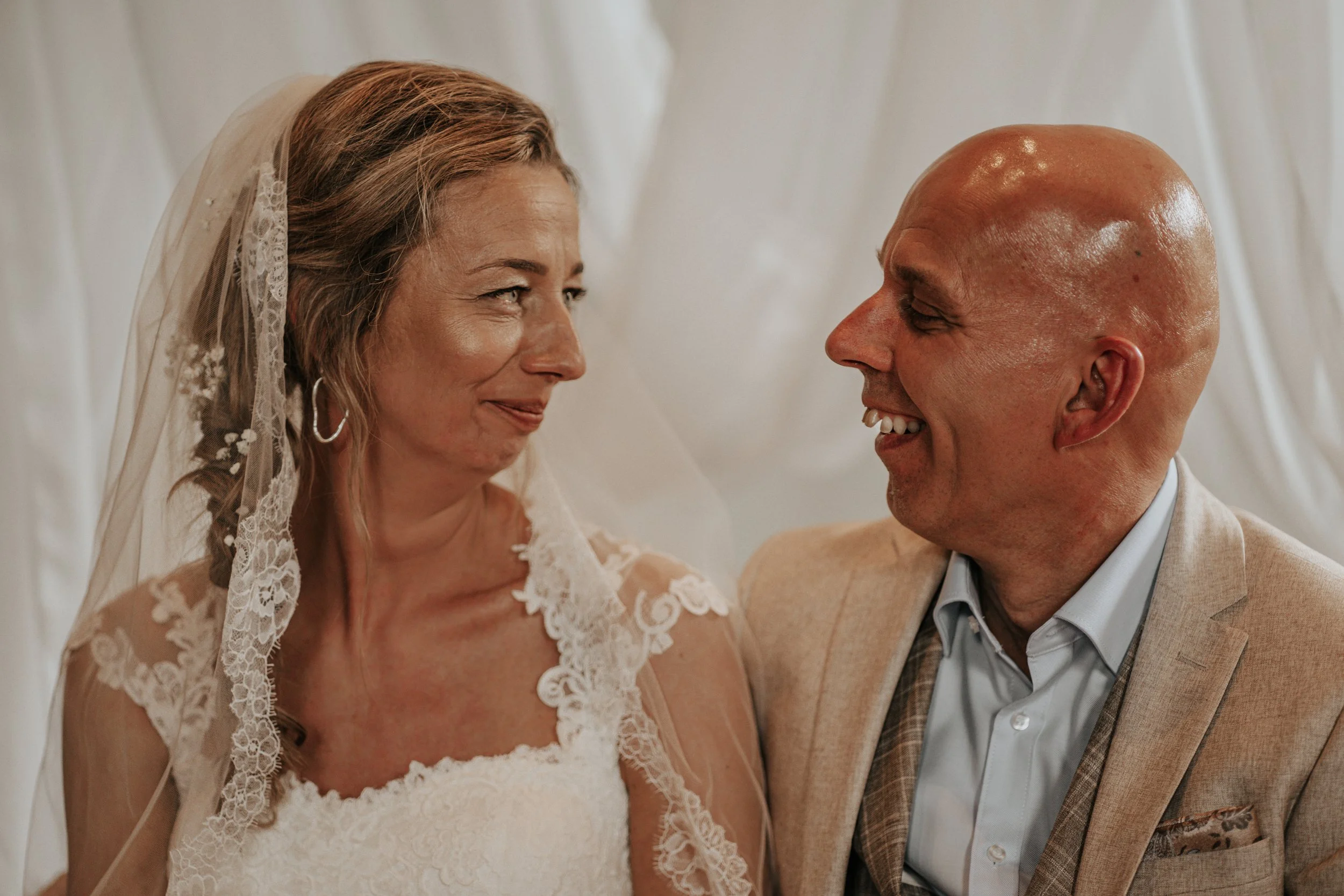 Een bruid en een bruidegom kijken elkaar glimlachend aan tijdens hun bruiloft, met een witte achtergrond.
