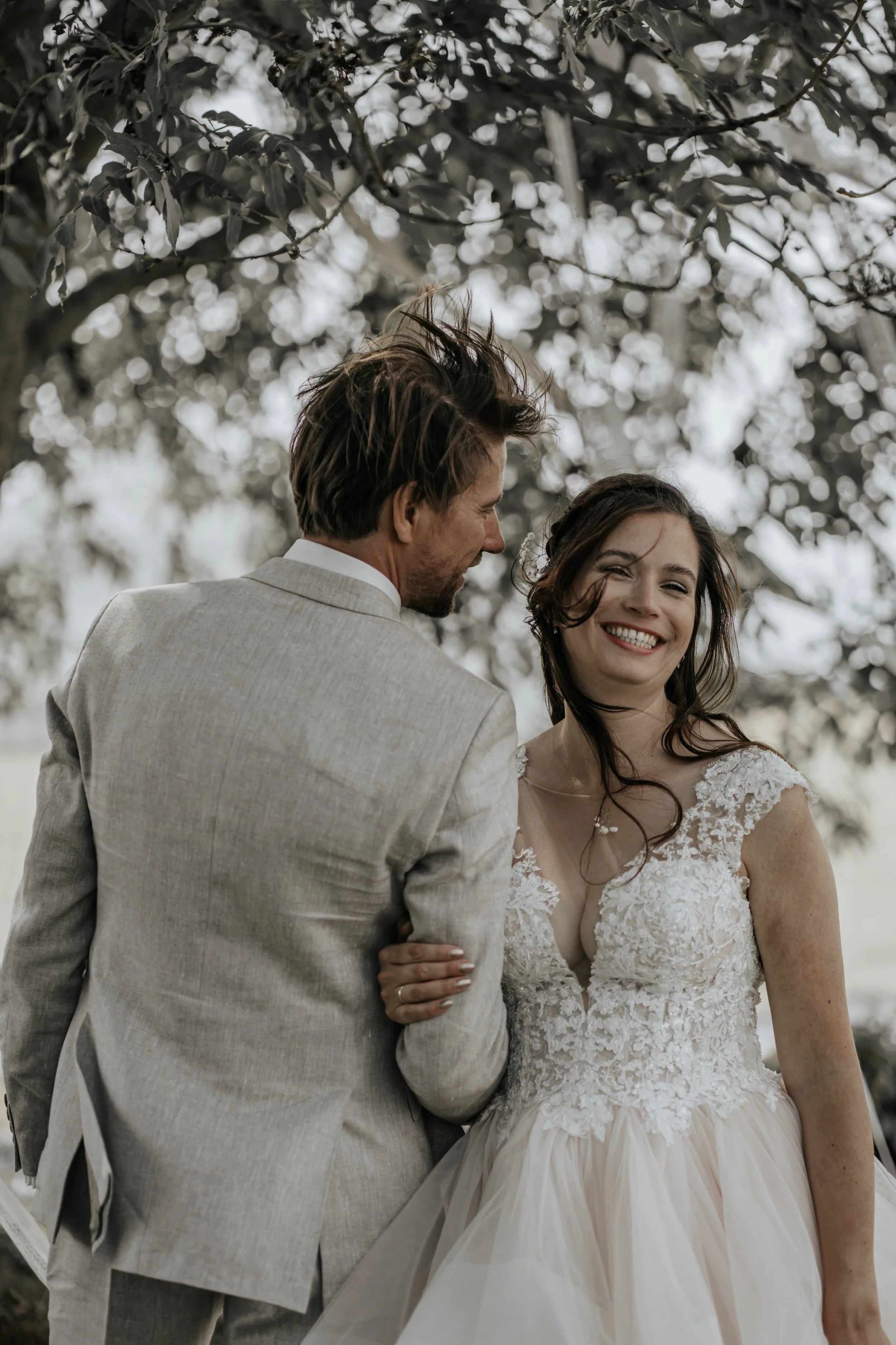 Een glimlachende vrouw in een witte trouwjurk naast een man in een lichtgrijze pak buiten onder een boom