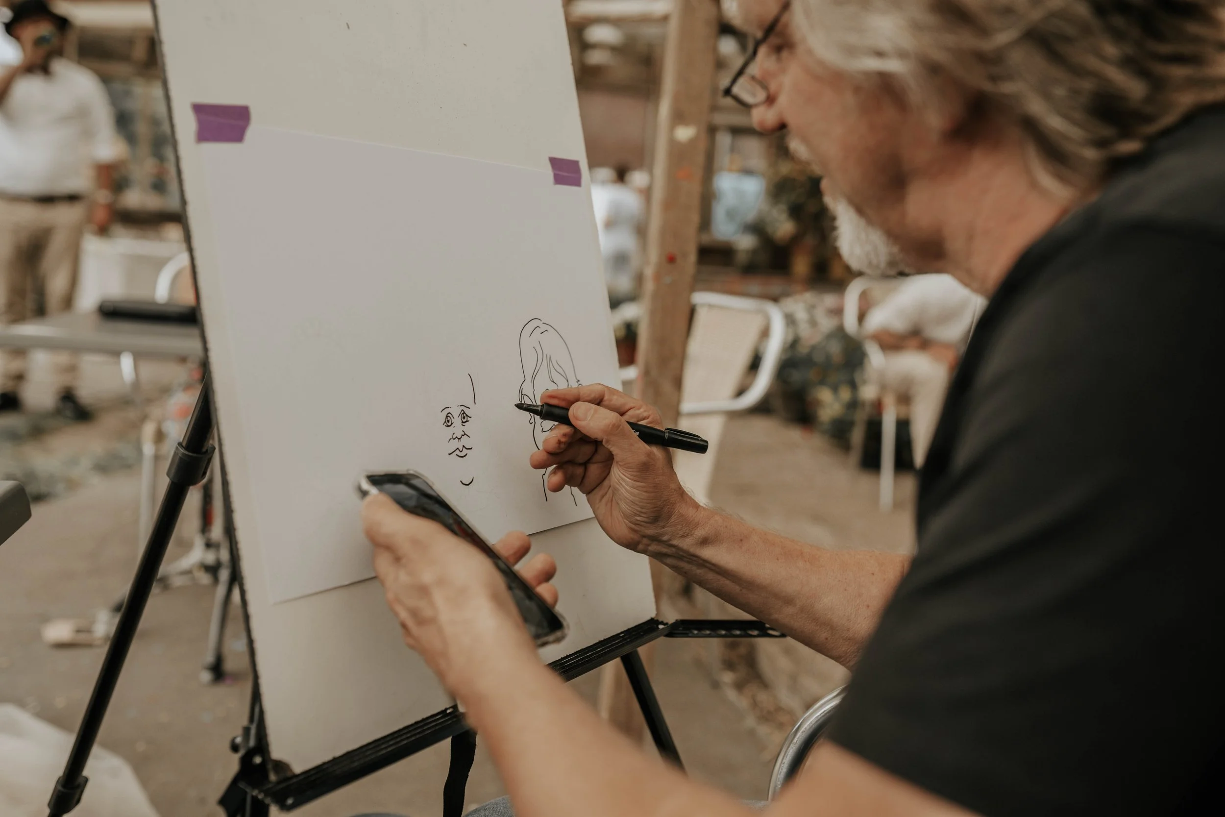 Een man die een tekening maakt op een wit bord terwijl hij een smartphone vasthoudt in een atelier of workshopruimte.