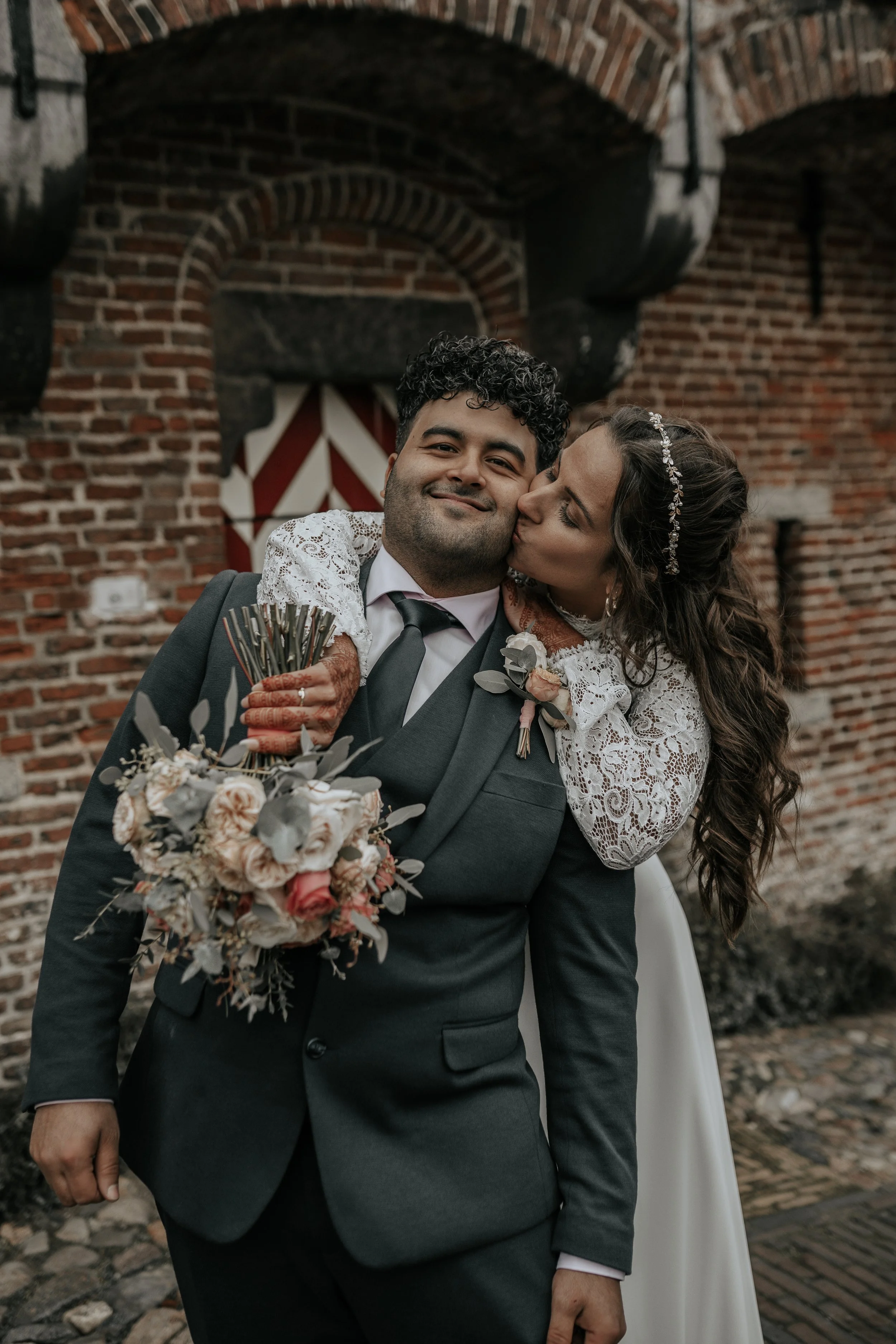 Een getrouwd paar op huwelijksdag, de vrouw kust de man op de wang, ze staan voor een bakstenen muur.