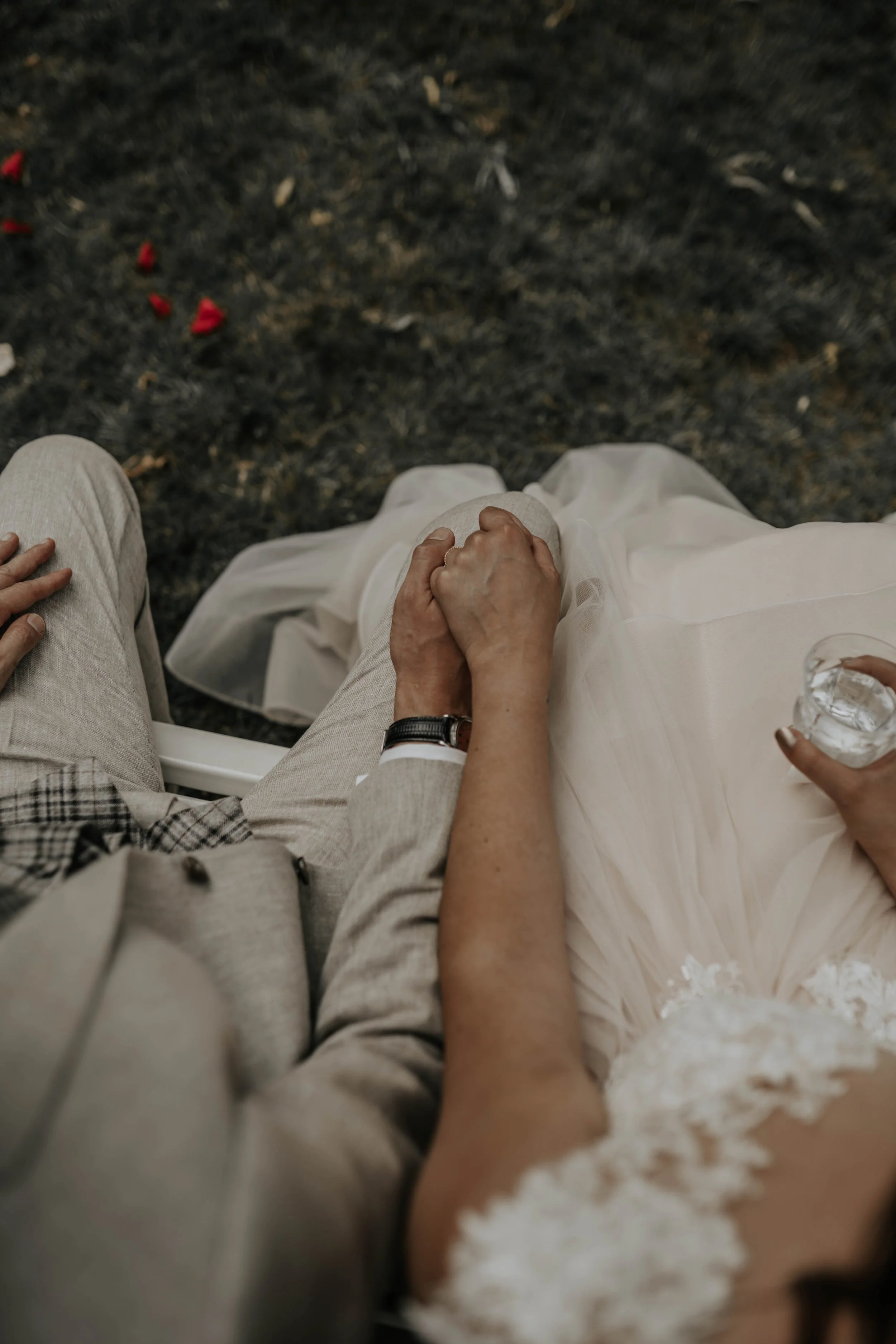 Close-up van een paar dat hand in hand zit, met de vrouw in een witte jurk en de man in een lichtkleurig pak, zittend op een bank op een donkere ondergrond.