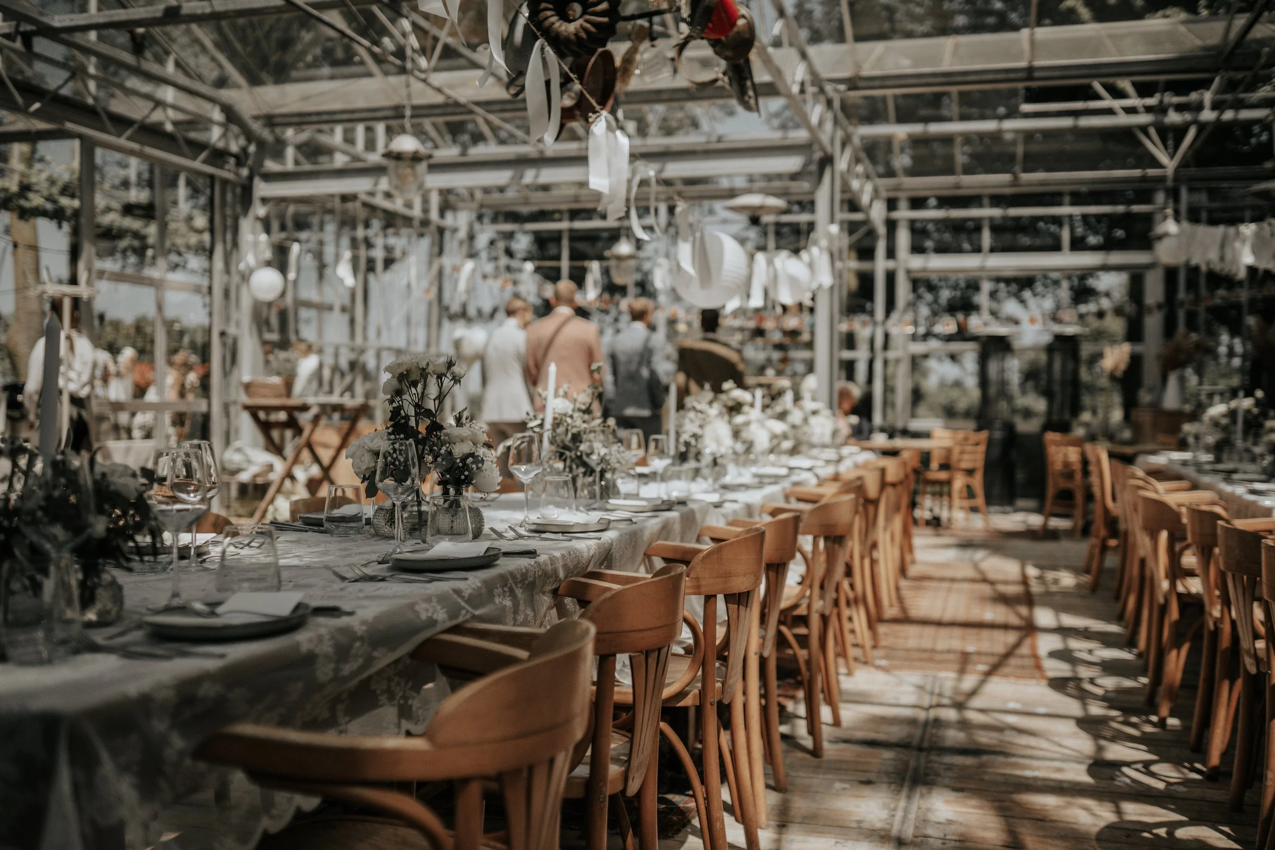 Feestelijke tafel in een serre met bloemen en gedekte borden, mensen op de achtergrond tijdens een bruiloft of feestje.