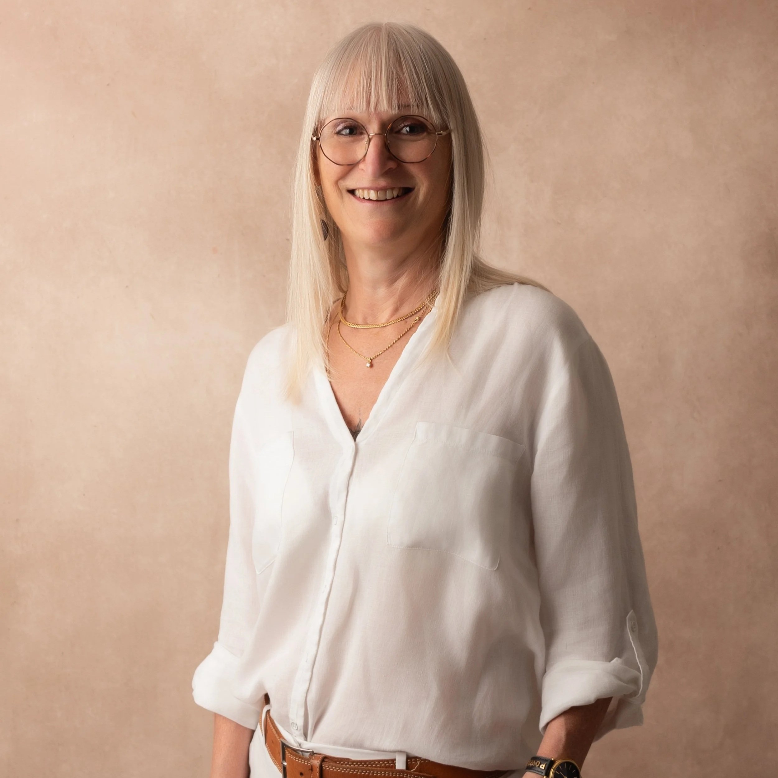 Une femme souriante avec des cheveux blonds et lunettes, portant une chemise blanche et des bijoux, posant devant un fond beige.
