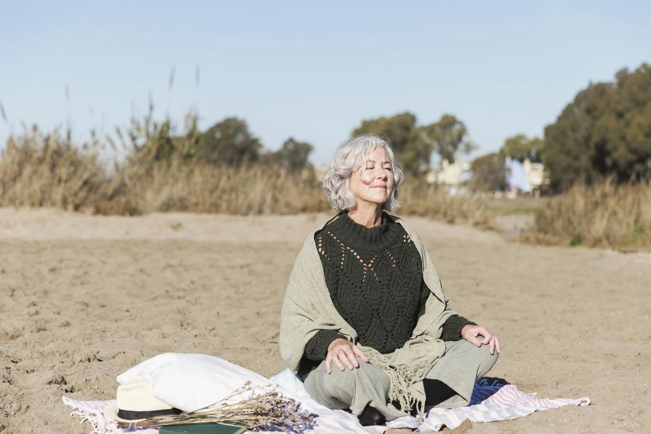 Une femme âgée assise en méditation sur une plage, en position de lotus, avec des yeux fermés et un visage paisible, entourée de sable, de végétation en arrière-plan, sous un ciel bleu clair.