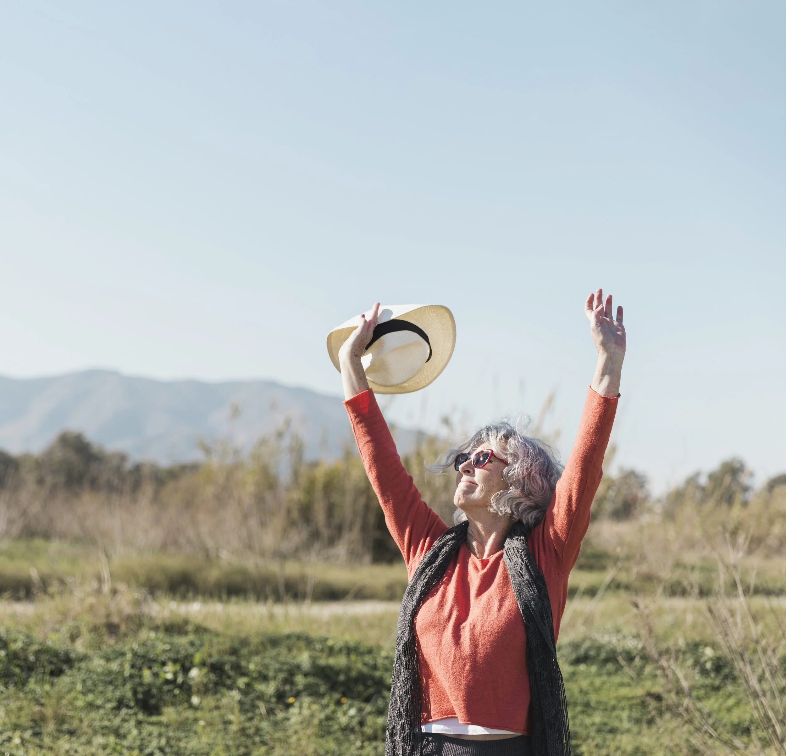 Une femme âgée avec des cheveux gris, portant des lunettes de soleil et un pull orange, lève les bras en l'air en tenant un chapeau de paille dans un paysage naturel avec des montagnes au loin et un ciel bleu clair.