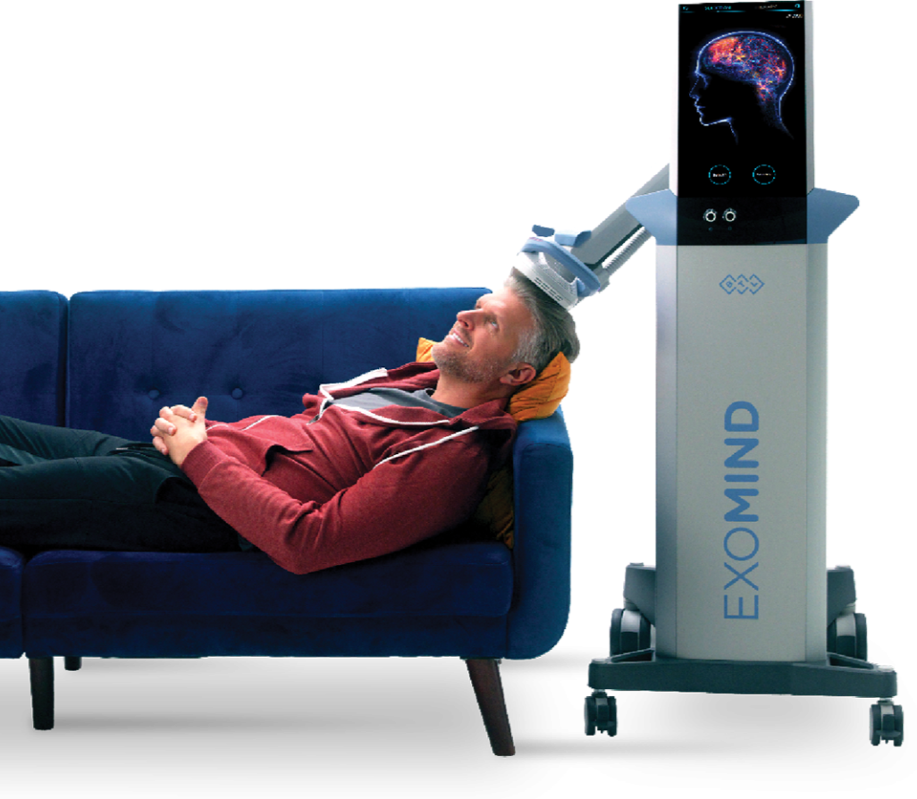 A man lying on a blue sofa with a yellow pillow, receiving a brain stimulation treatment from a medical device labeled 'EXOMIND', with a screen showing a digital representation of a human brain and a profile of a person's head