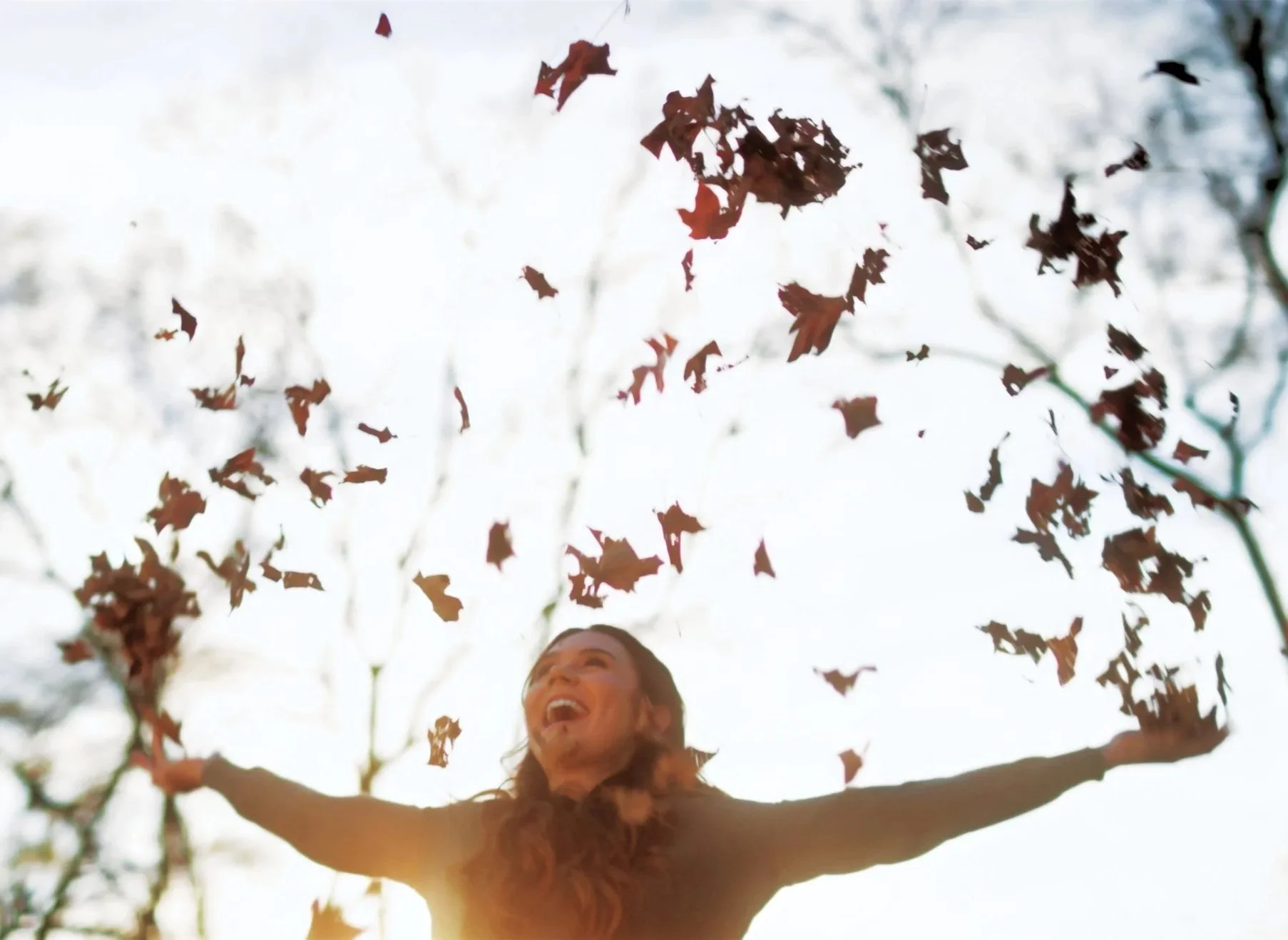 A woman with long hair joyfully throwing fallen leaves into the air outdoors on a sunny day.