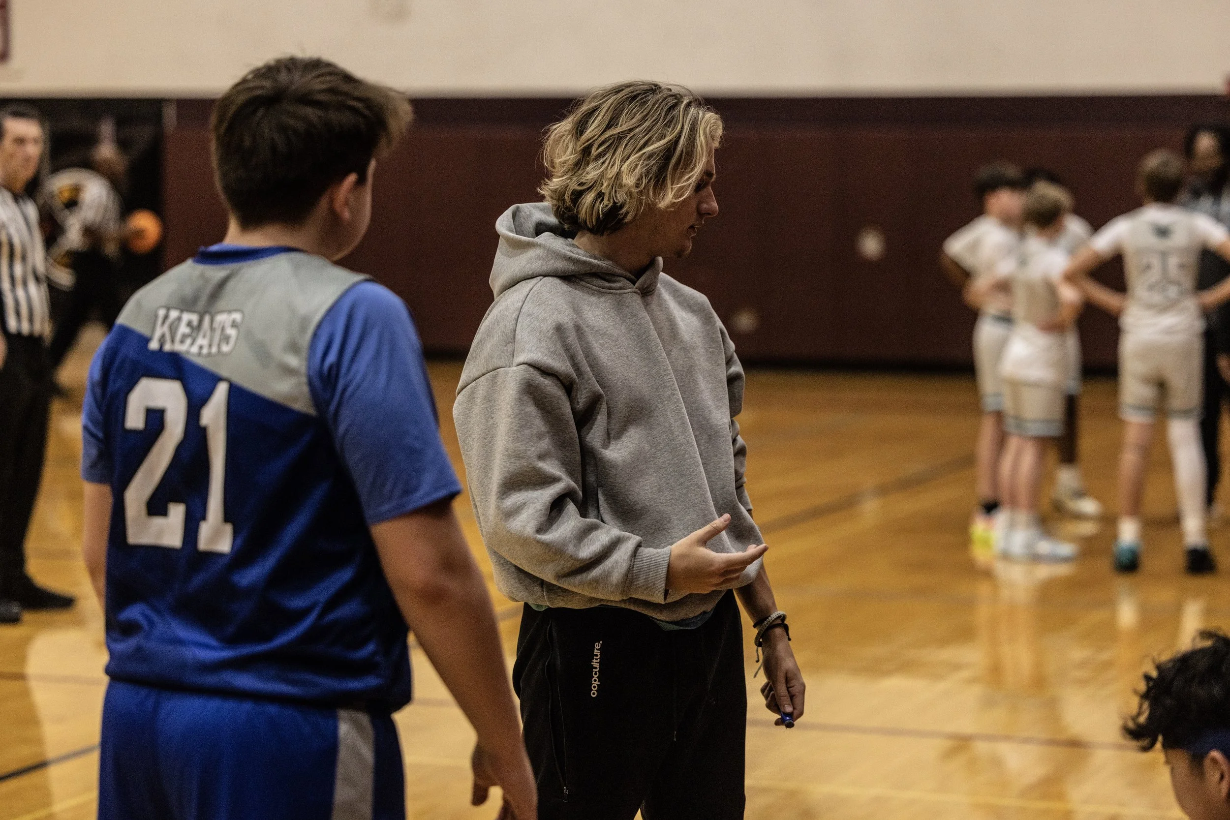 A coach or adult standing on a gymnasium floor talking to a young male athlete wearing a blue jersey with number 21. In the background, other young athletes in white sports uniforms are gathered in a group.