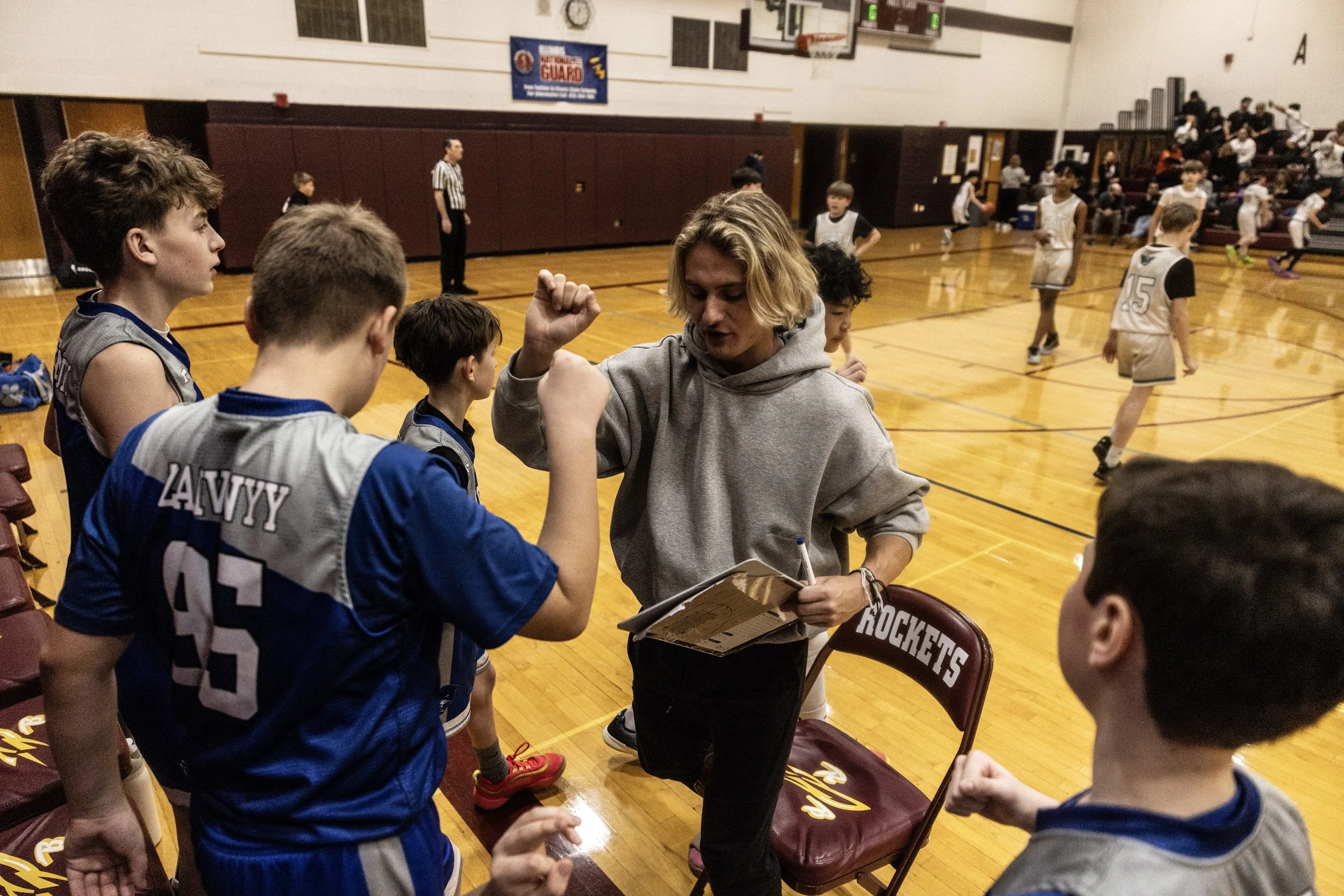 A female coach in a gray hoodie giving motivational advice to young male basketball players on the bench during a game in an indoor gymnasium.