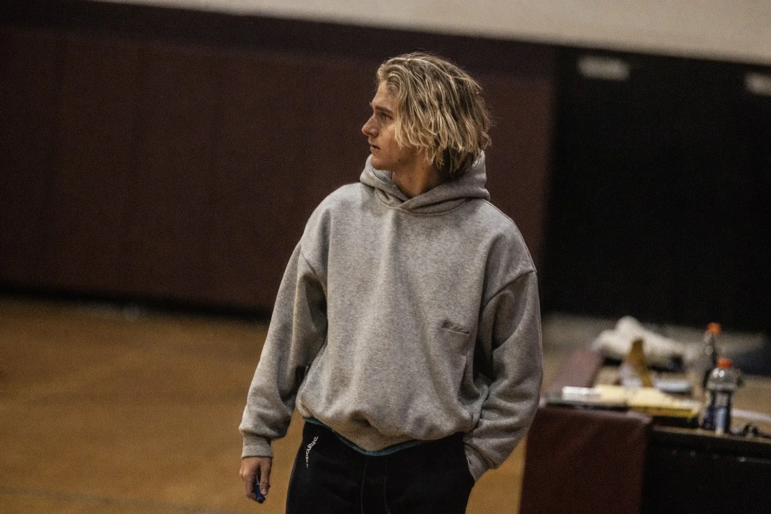 A young man with curly blond hair wearing a gray hoodie standing in a room with a brown wall and a table with various items on it.