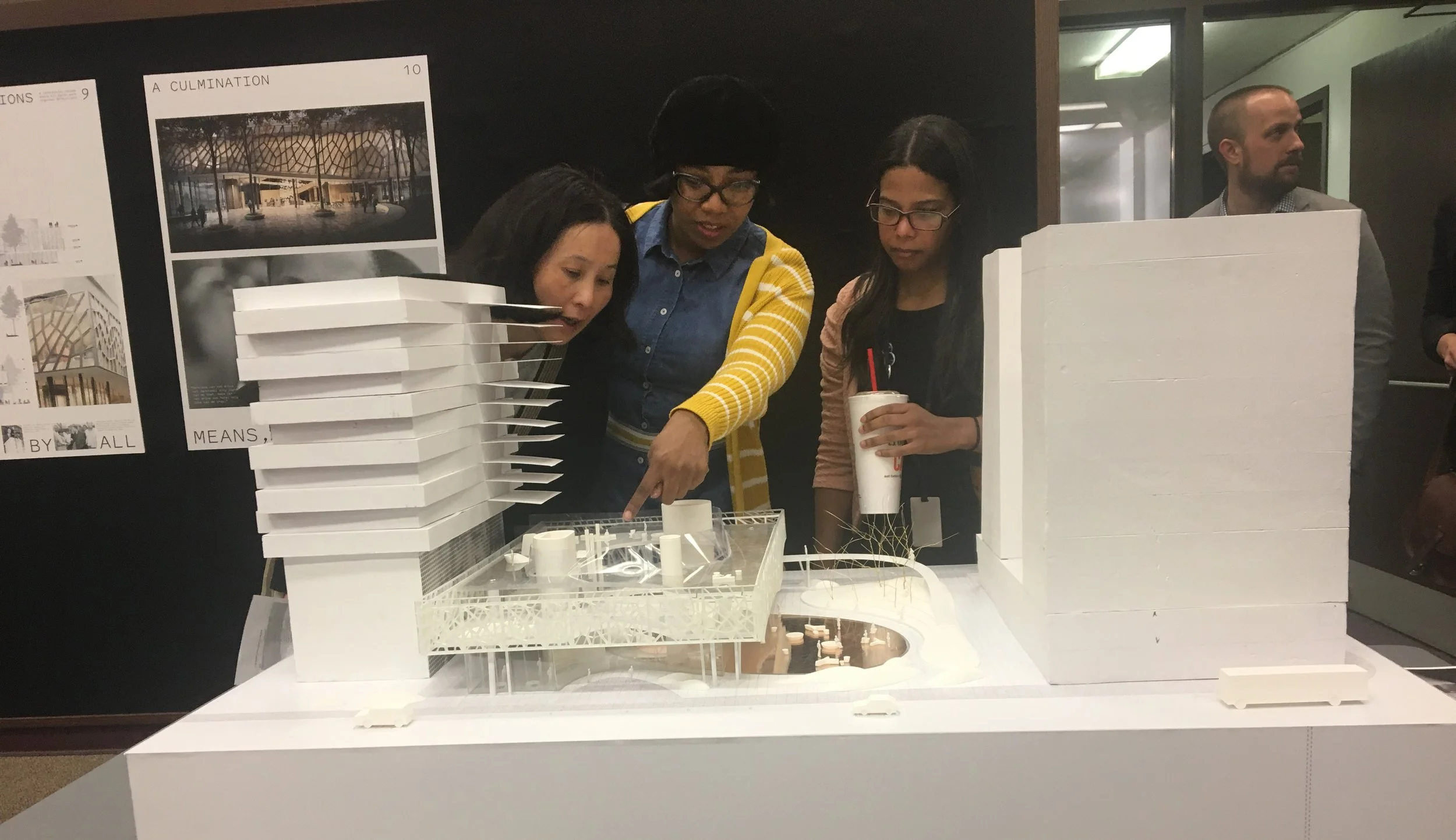 Three women and one man looking at an architectural model of a building on display, with poster boards in the background.