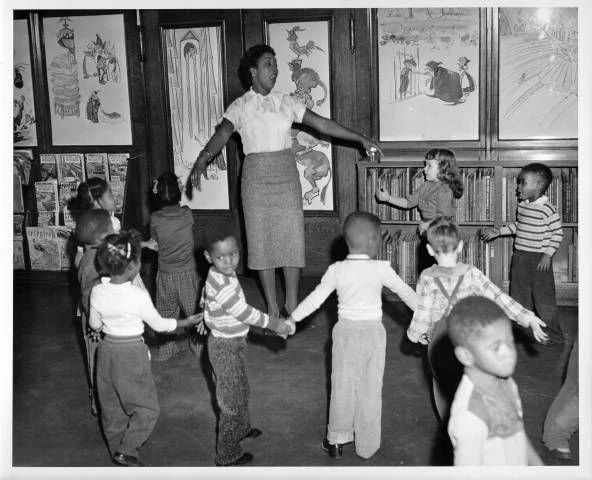 A librarian in Cleveland Ohio leading a circle game with young children in a classroom, with children's artwork displayed on the walls.
