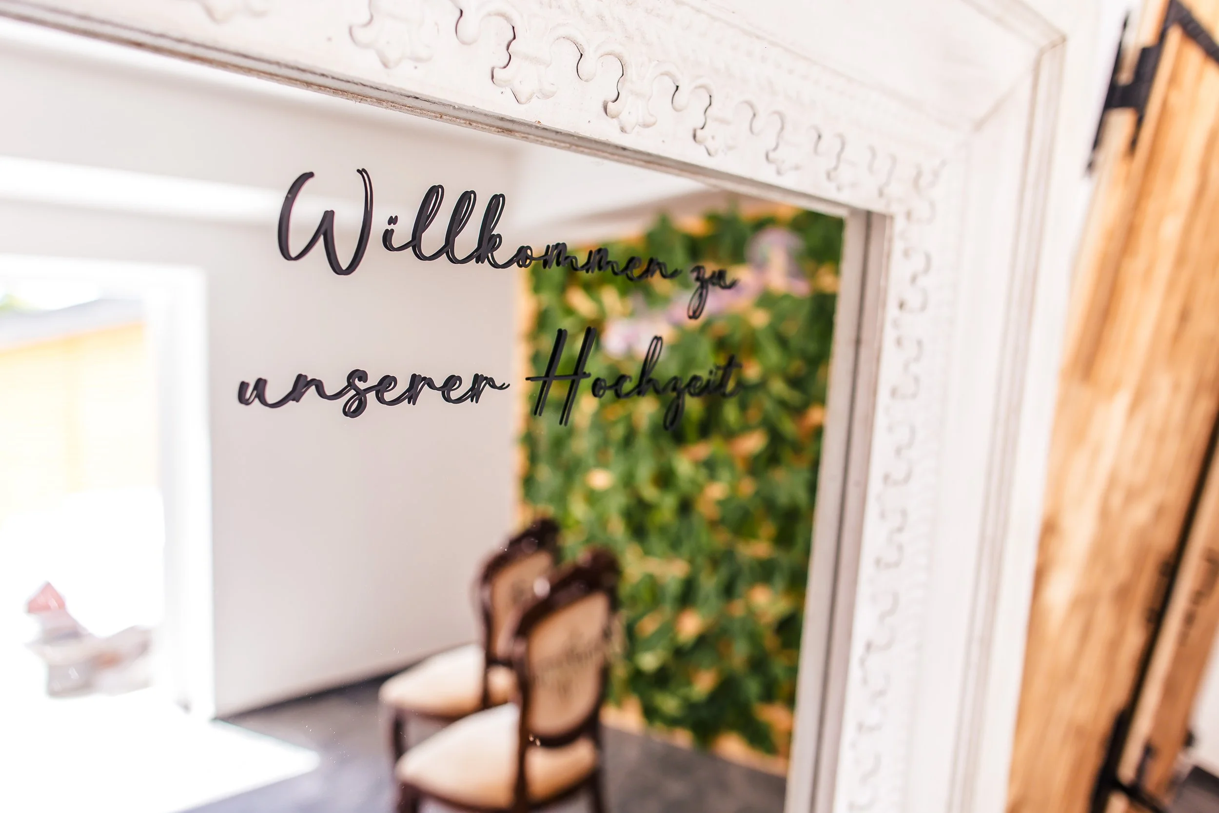 Spiegel mit der Aufschrift 'Willkommen zur unserer Hochzeit', dahinter sind Bänke und ein grüner Baum sichtbar.