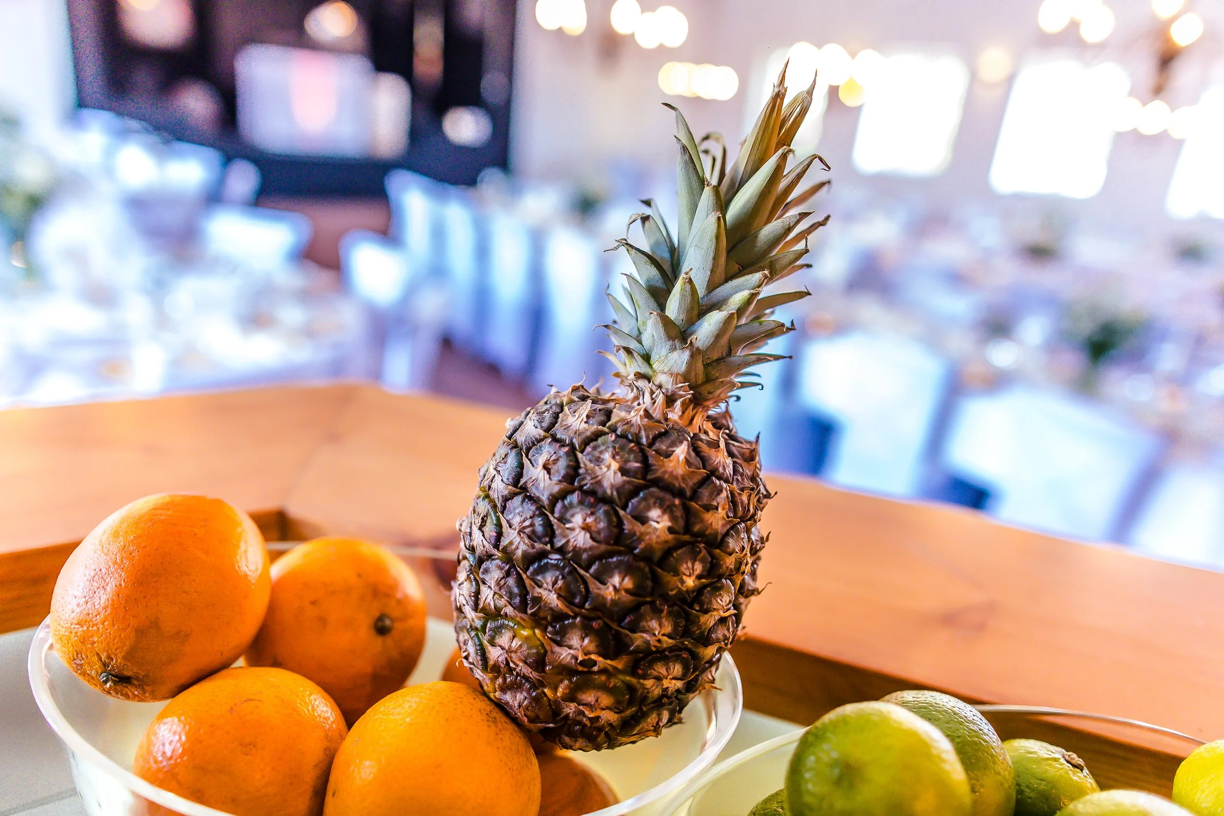 Eine Ananas auf einem Teller neben Orangen und Limetten, im Hintergrund ein Esszimmer mit decorativen Lichtern.