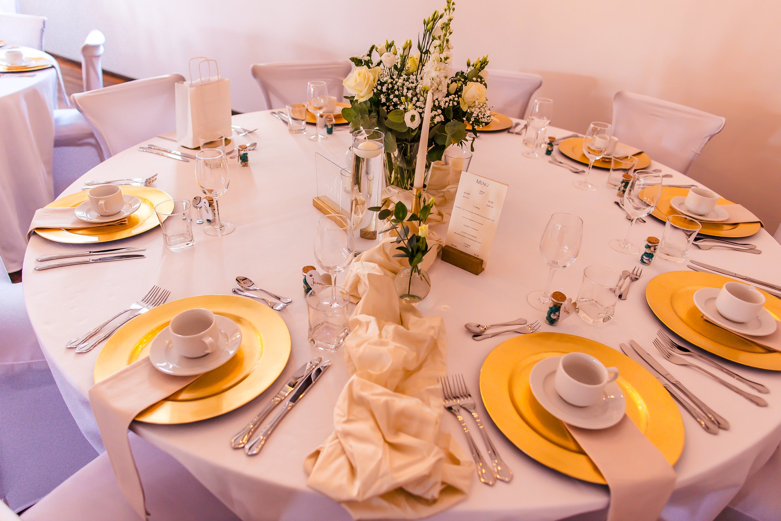 Ein festlich gedeckter runder Esstisch mit weißen Tischdecken, goldenen Tellern, weißen Tassen, Gläsern und Besteck, dekoriert mit einer großen Blumenmitte in Vasen und Kerzen, für eine Feier oder ein Abendessen vorbereitet.