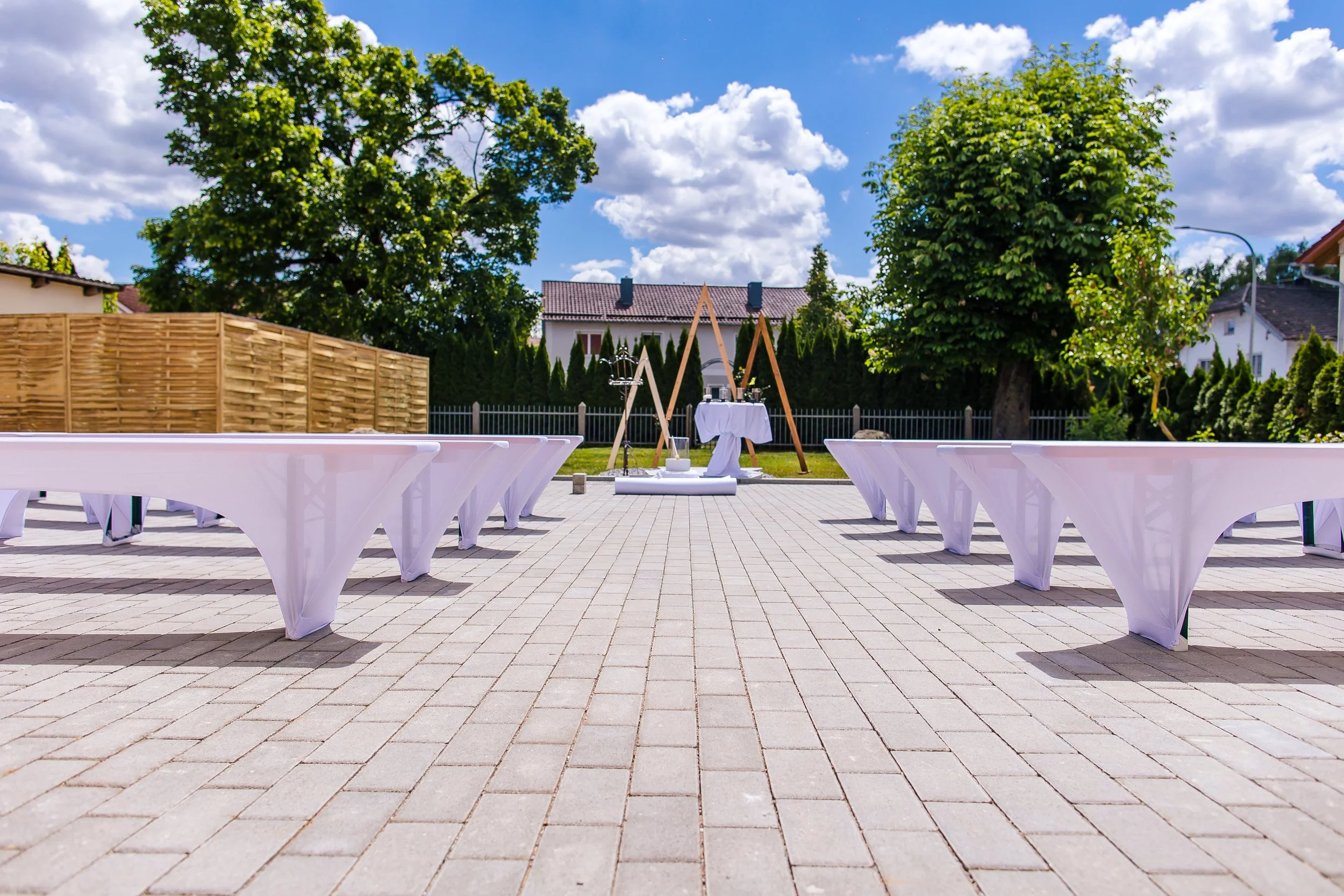 Ein im Freien aufgebauter Festort mit weißen Tischen, die mit weißen Tuch bedeckt sind, auf einem gepflasterten Platz. Im Hintergrund sind Holzgestelle, eine kleine runde white Zelt und ein Tisch mit Dekorationen. Es ist sonnig mit blauen Himmel und 