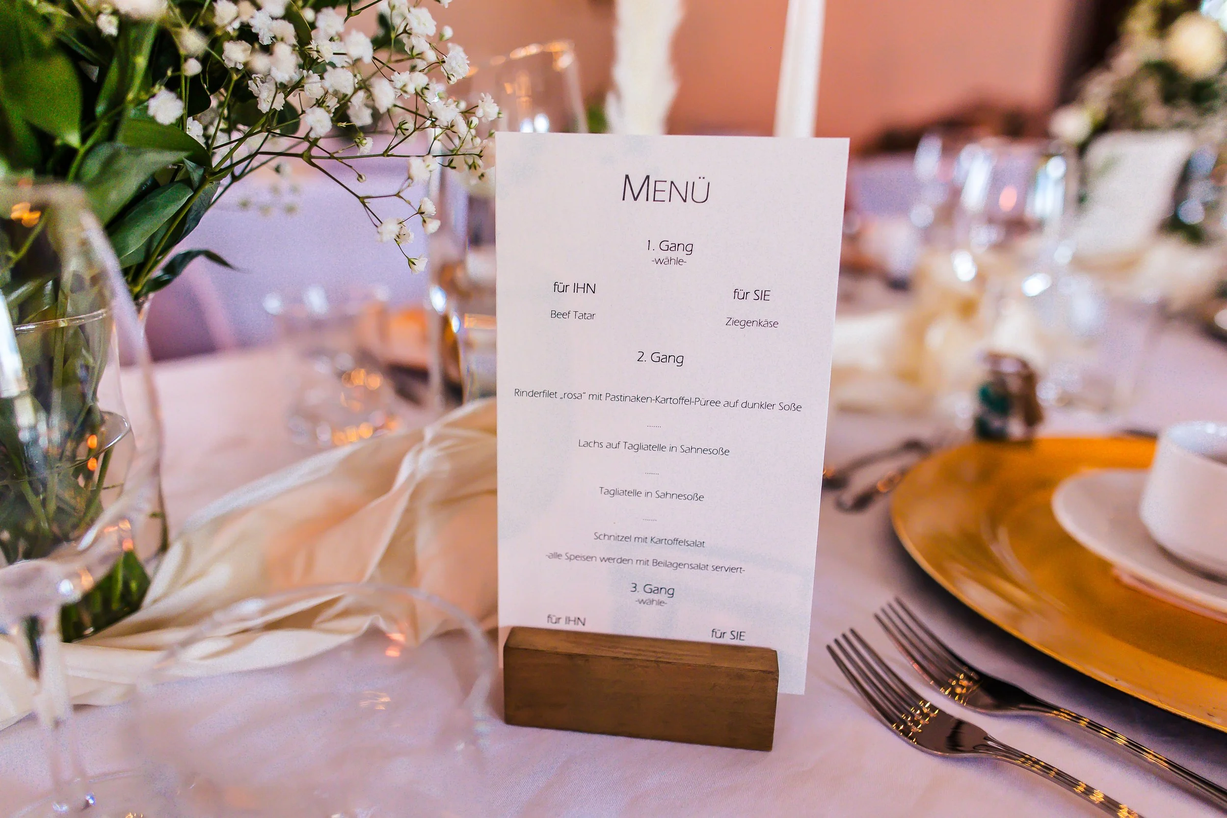 Speisekarte auf einem Hochzeitstisch, umgeben von Blumen, Gläsern und Tellern.