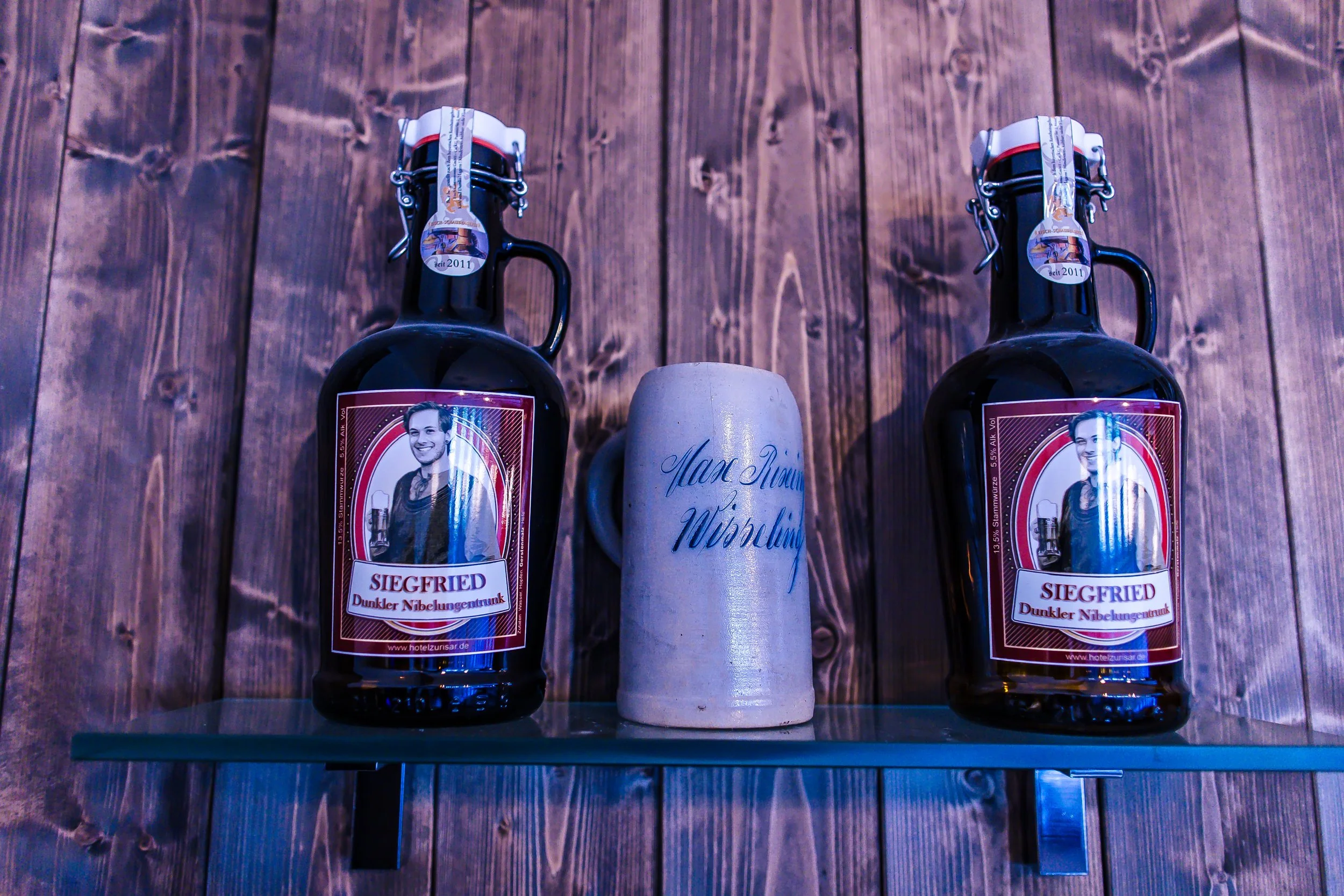 Zwei dunkle Bierflaschen mit Etiketten und ein Bierkrug, der auf einem Glasregal an einer Holzvertäfelung steht.