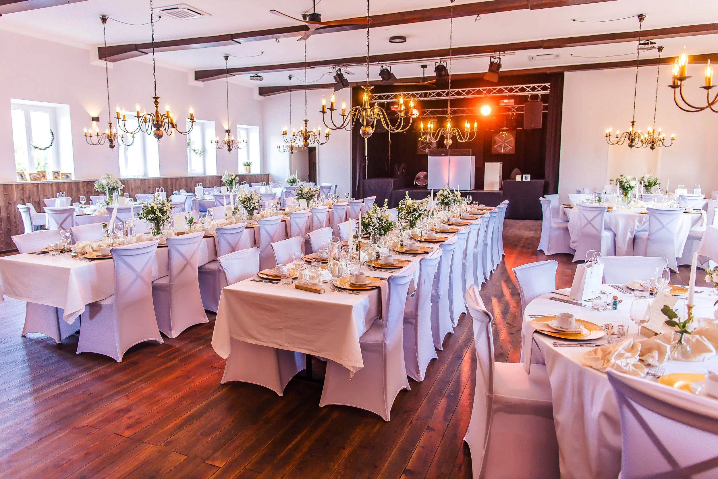 Festlich gedeckter Veranstaltungsraum mit weißen Tischen und Stühlen, Kerzen, Blumen und einer kleinen Bühne mit Lichttechnik.