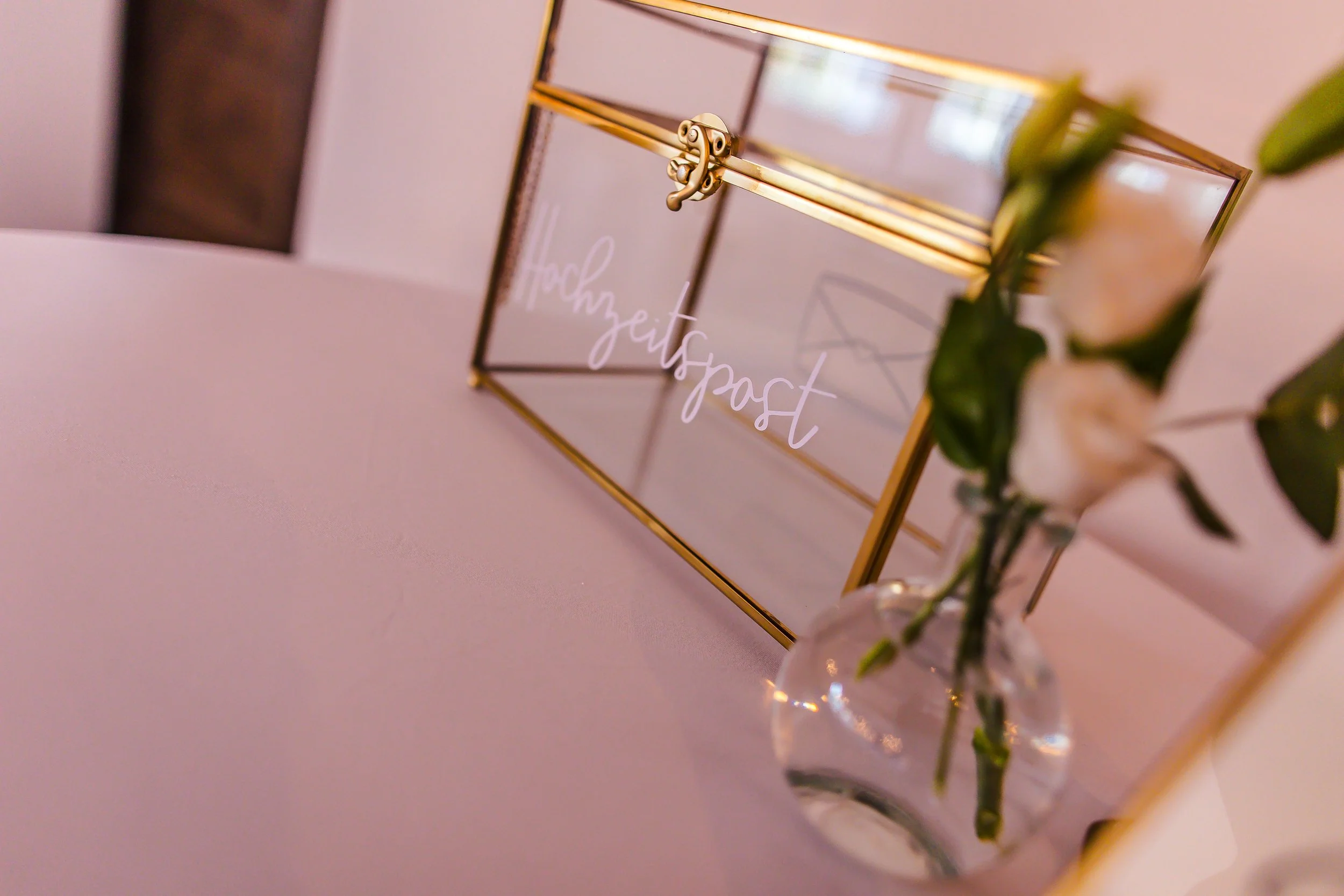 Eine goldene Box mit der Aufschrift „Hochzeit“ steht auf einem Tisch, im Vordergrund eine Vase mit weißen Blumen.
