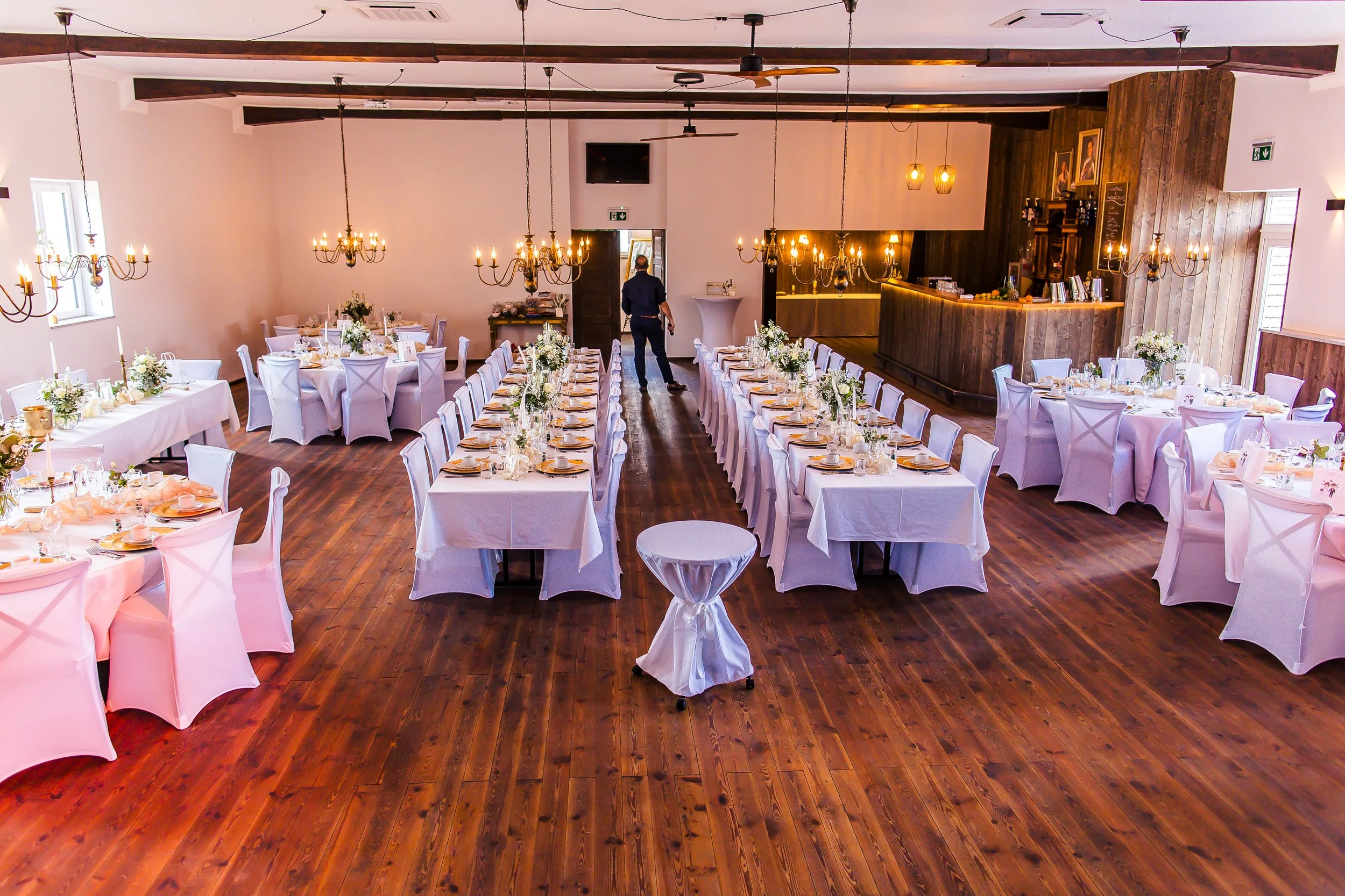 Ein festlich gedeckter Saal mit weißen Tischen und Stühlen, Blumenarrangements, Kerzen und Lichterketten, bereit für eine Feier.