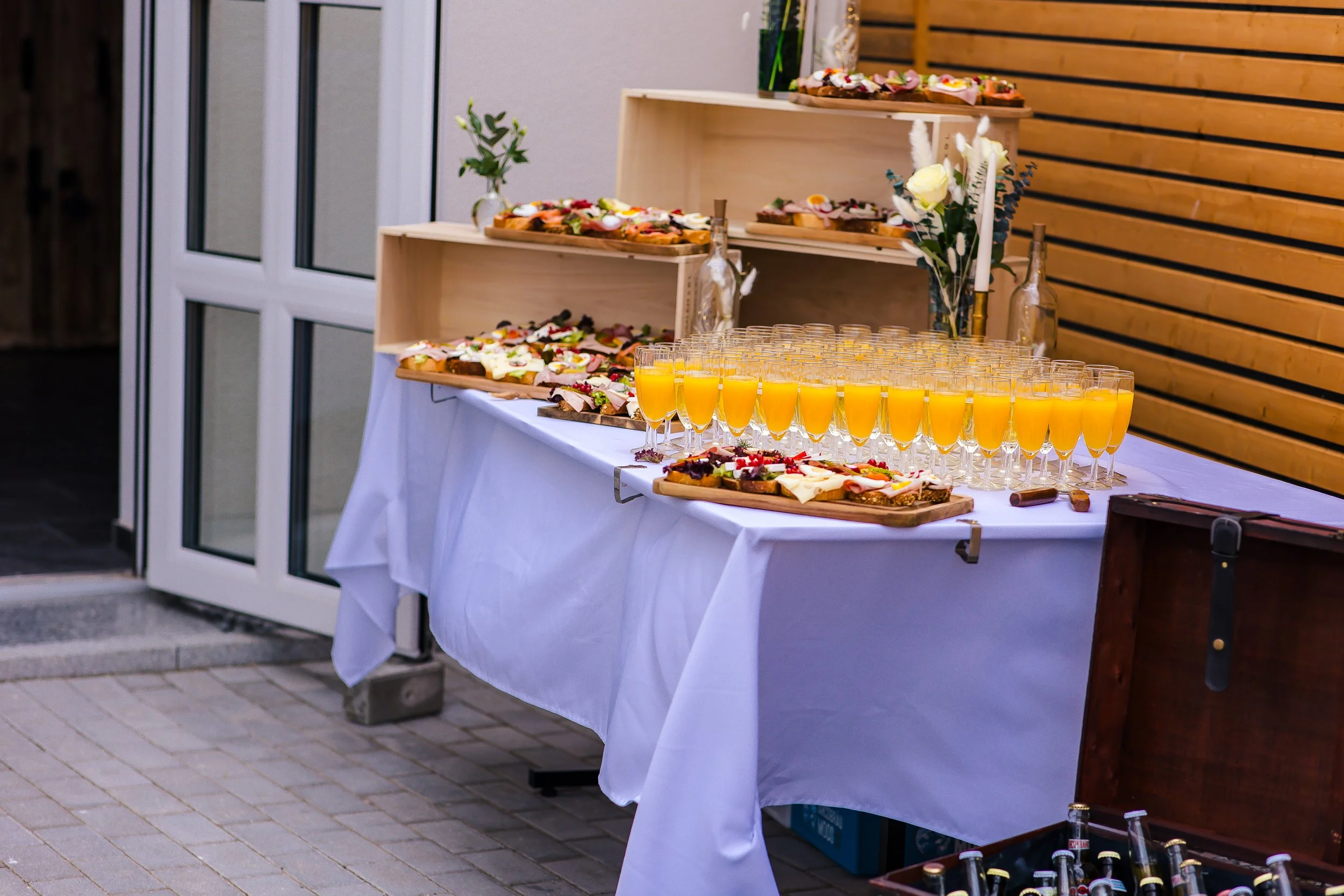 Buffet-Tisch mit verschiedenen belegten Broten, Gläsern mit Orangensaft, Blumen und Kerzen, im Außenbereich bei Sonnenlicht.