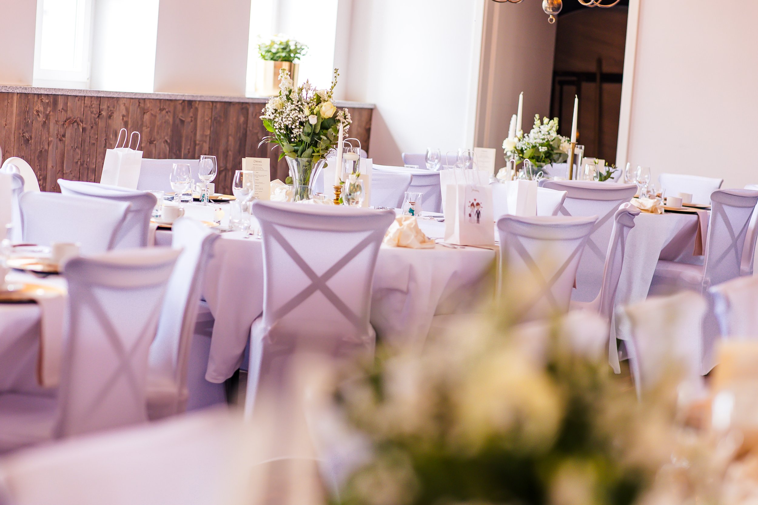 Festlich gedeckter Raum mit weißen Tischen, Stühlen und Blumenarrangements, vorbereitet für eine Veranstaltung.