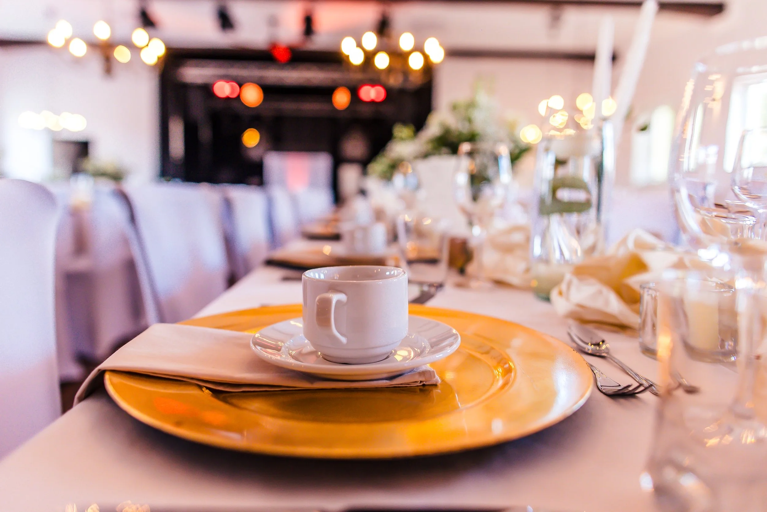 Ein schön gedeckter Tisch für eine Veranstaltung, mit einem weißen Kaffeetasse auf goldenem Teller, Sitzordnung, Gläsern und Blumen, in einem eleganten Raum mit warmem Licht.