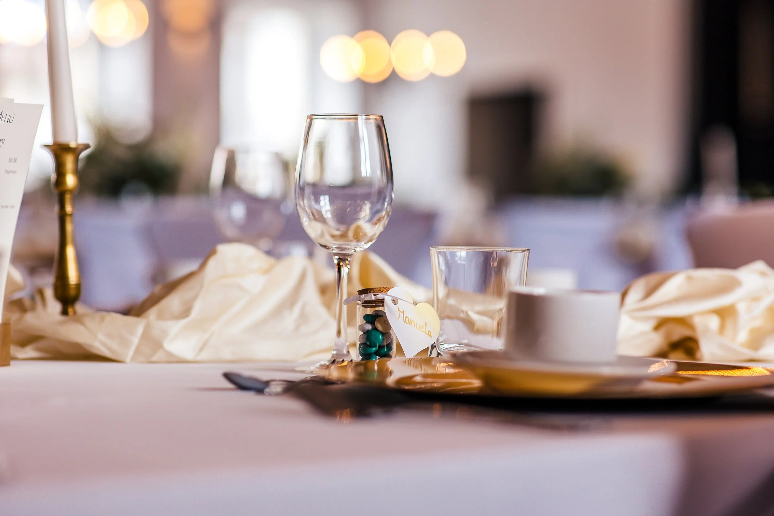 Ein gedeckter Tisch mit Weinglas, Tasse, Teelöffel, Kerze, Schnullerkette mit Herz, im Hintergrund diffuse Lichter.