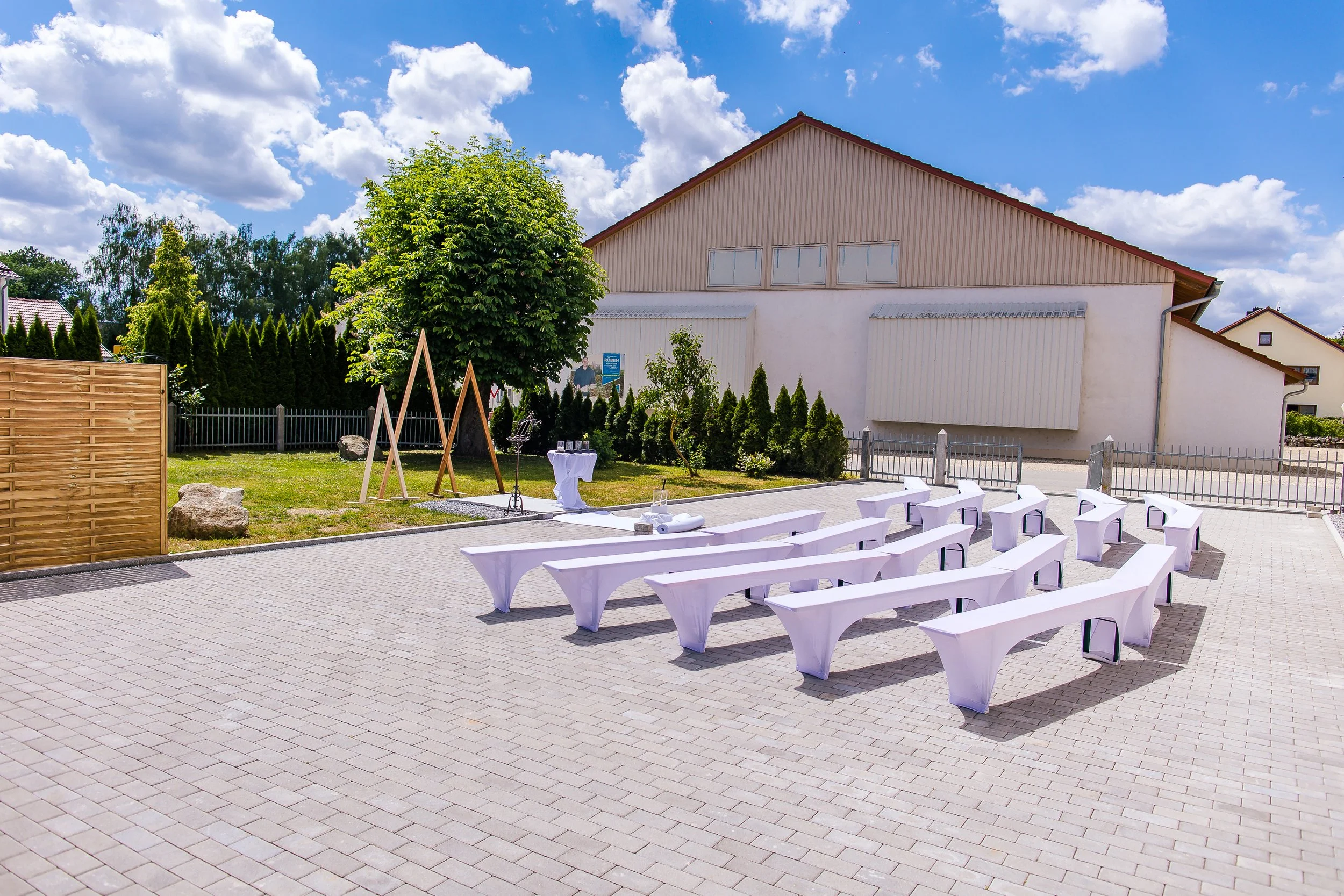 Außengelände mit weißen Bierzeltgarnituren für eine Veranstaltung, unbedeckter Himmel mit Wolken, grüner Baum, Gartenbereich und Gebäude im Hintergrund.