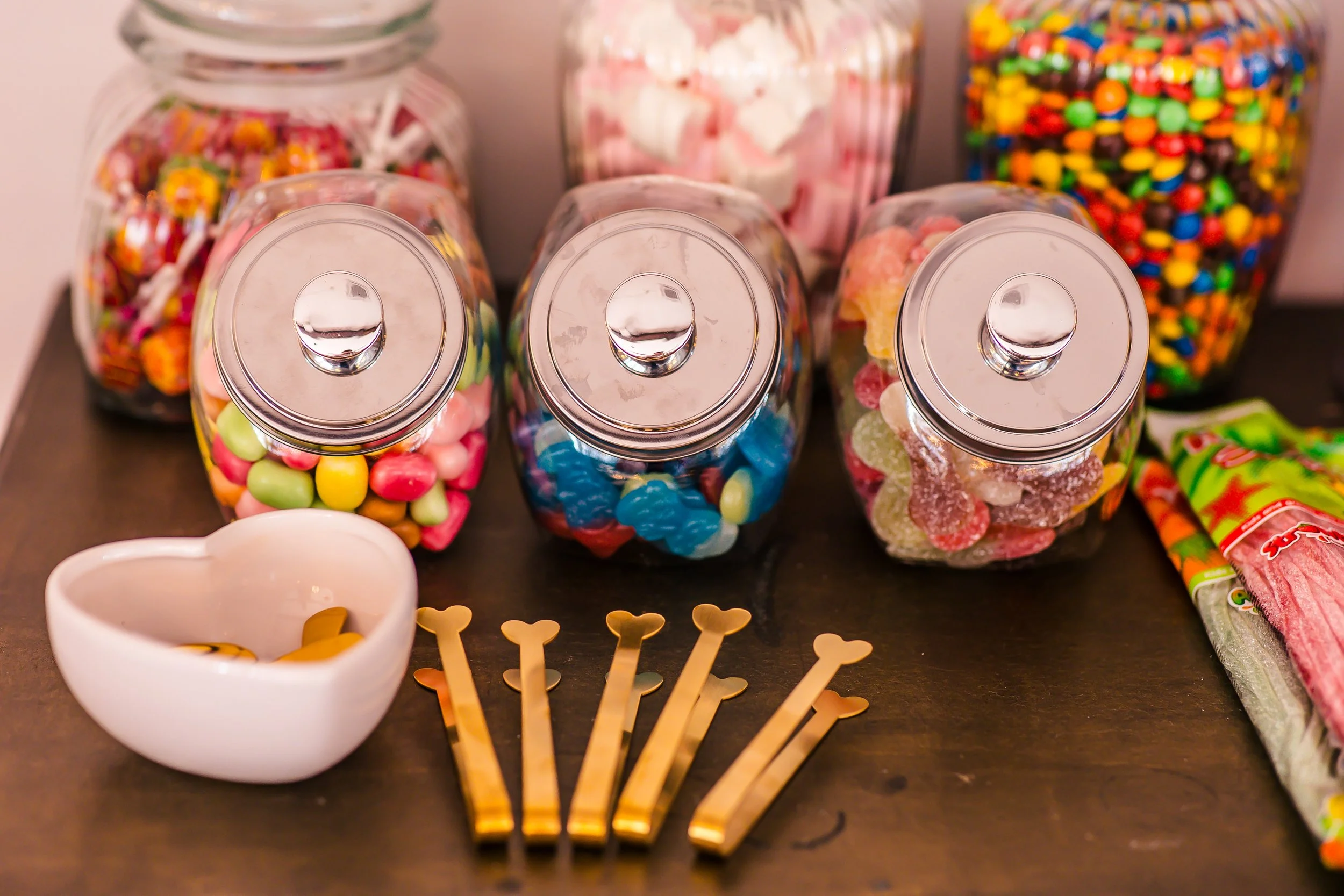 bunte Süßigkeiten in Glasvasen, dazu ein weißes Schälchen mit Bonbons und goldene Löffel auf einem Tisch.